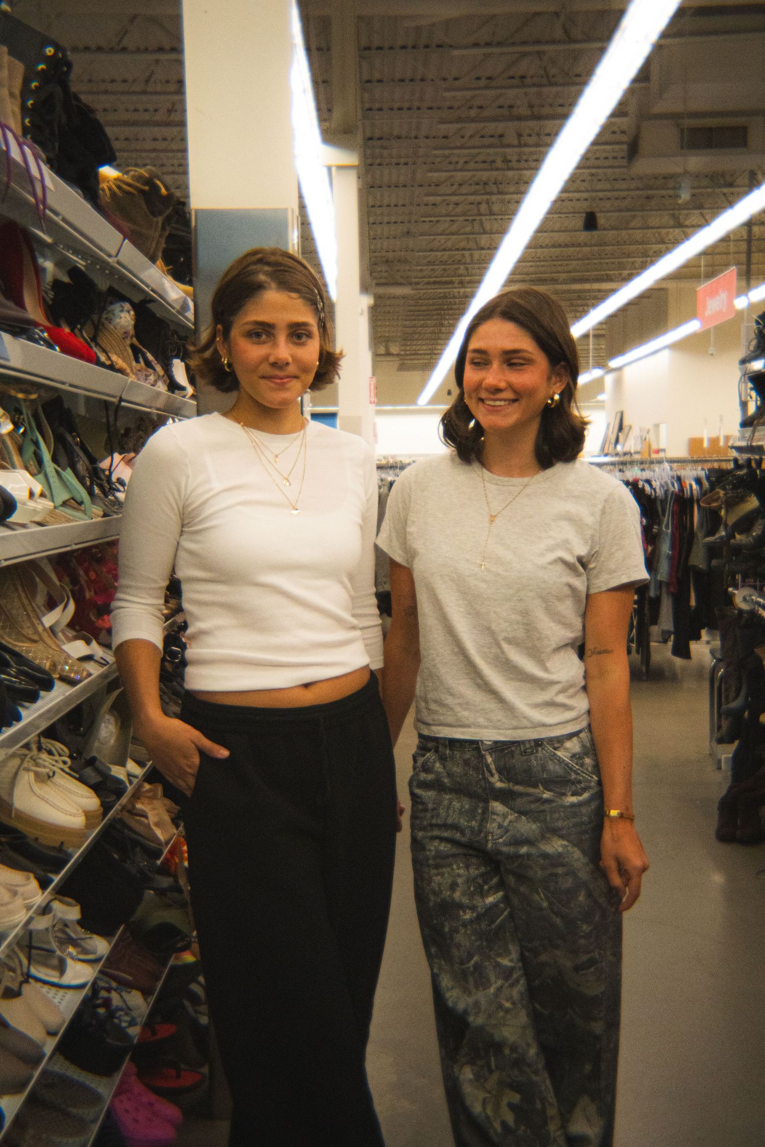 Two smiling women in casual white tops pose in a thrift store shoe department.