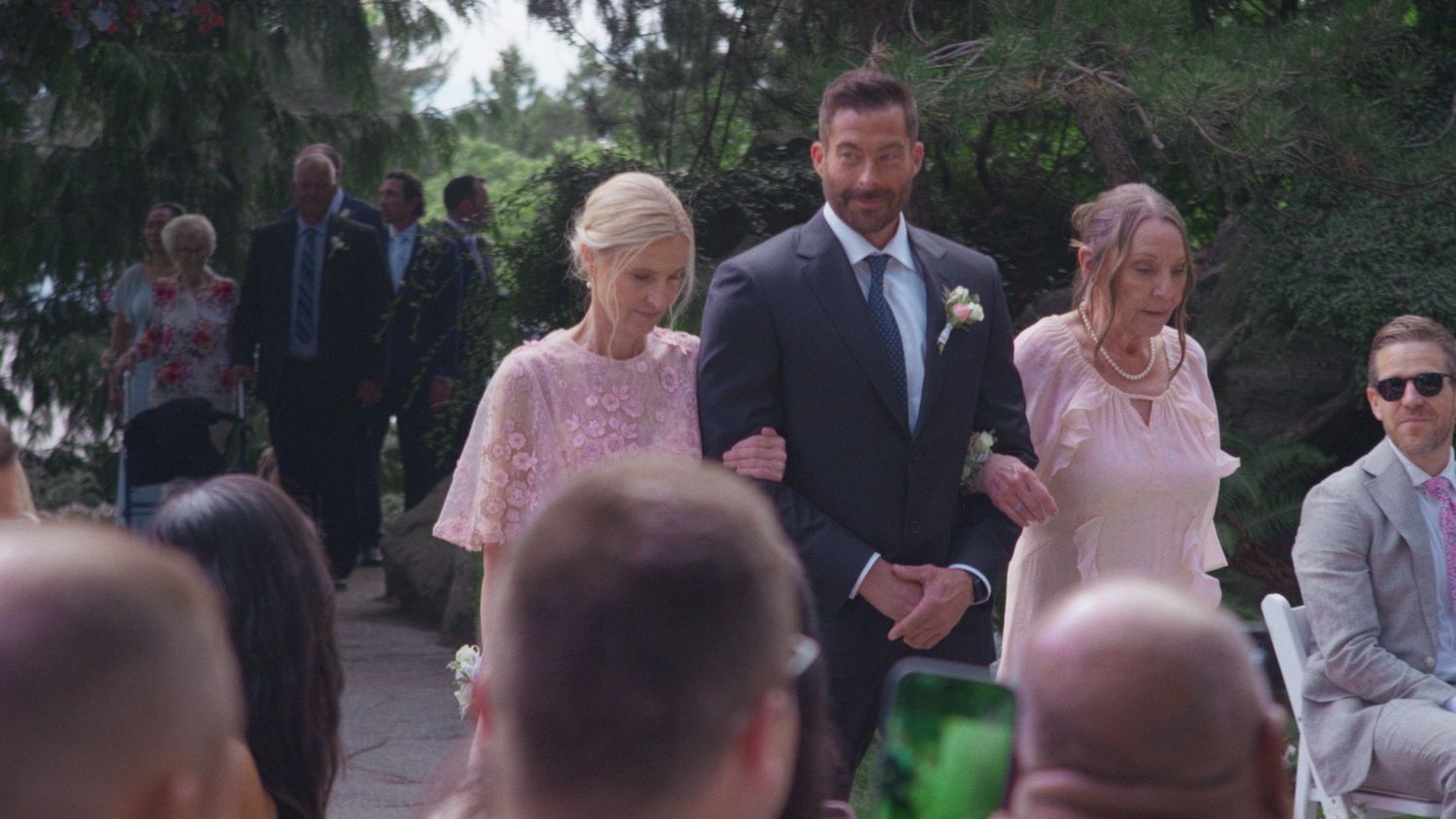 A man in a navy suit escorts two women in pink dresses down a wedding aisle outdoors.