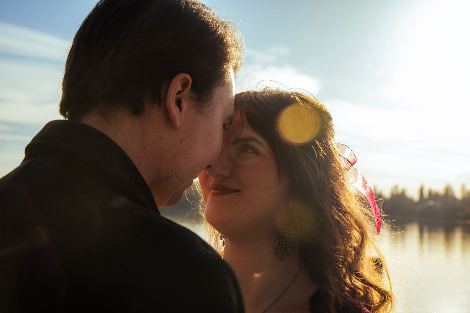 Romantic couple sharing an intimate moment nose-to-nose with bright sunlight and a lake shimmering behind them.