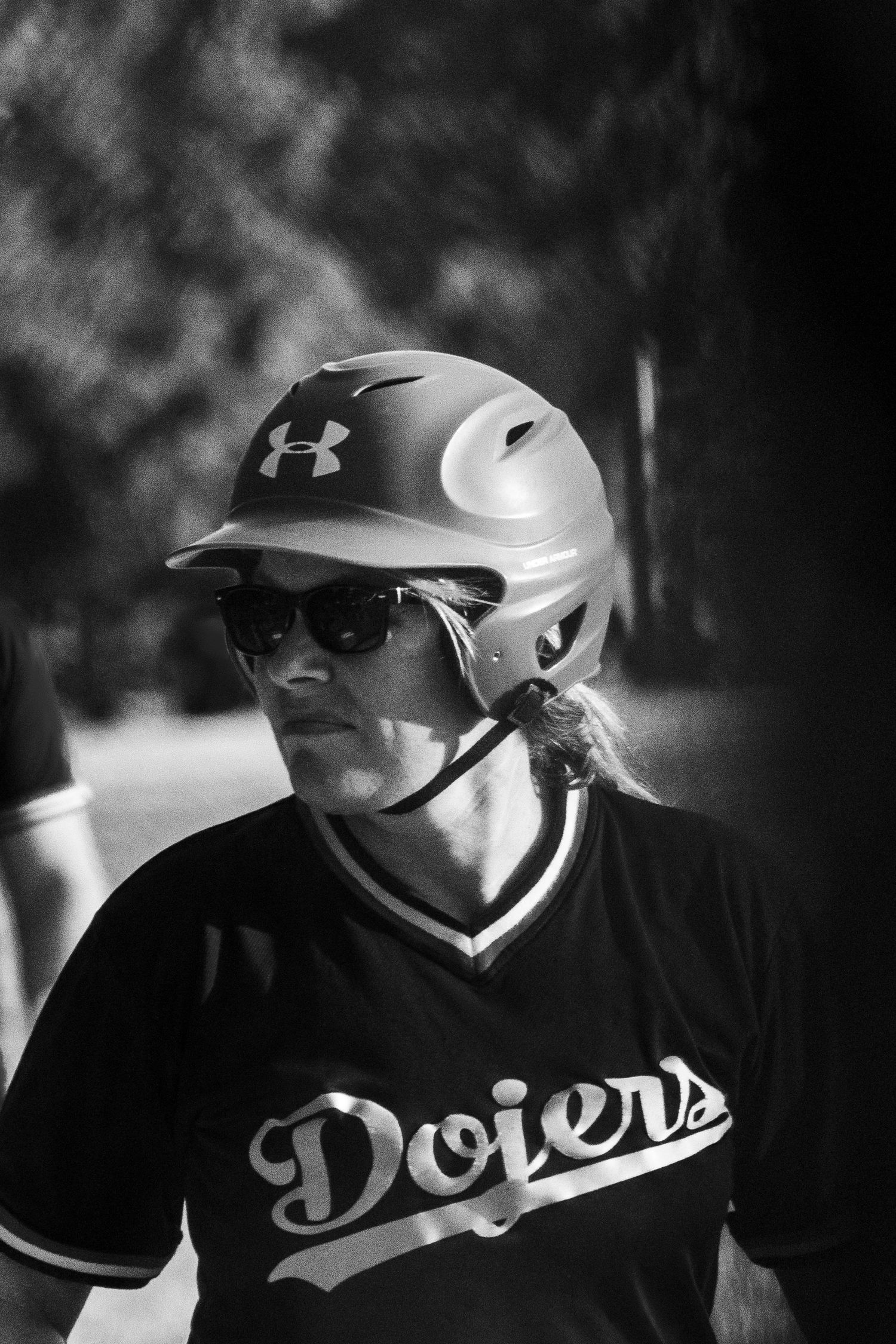 Female softball player wearing an Under Armour helmet and Dodgers jersey with sunglasses in black and white.