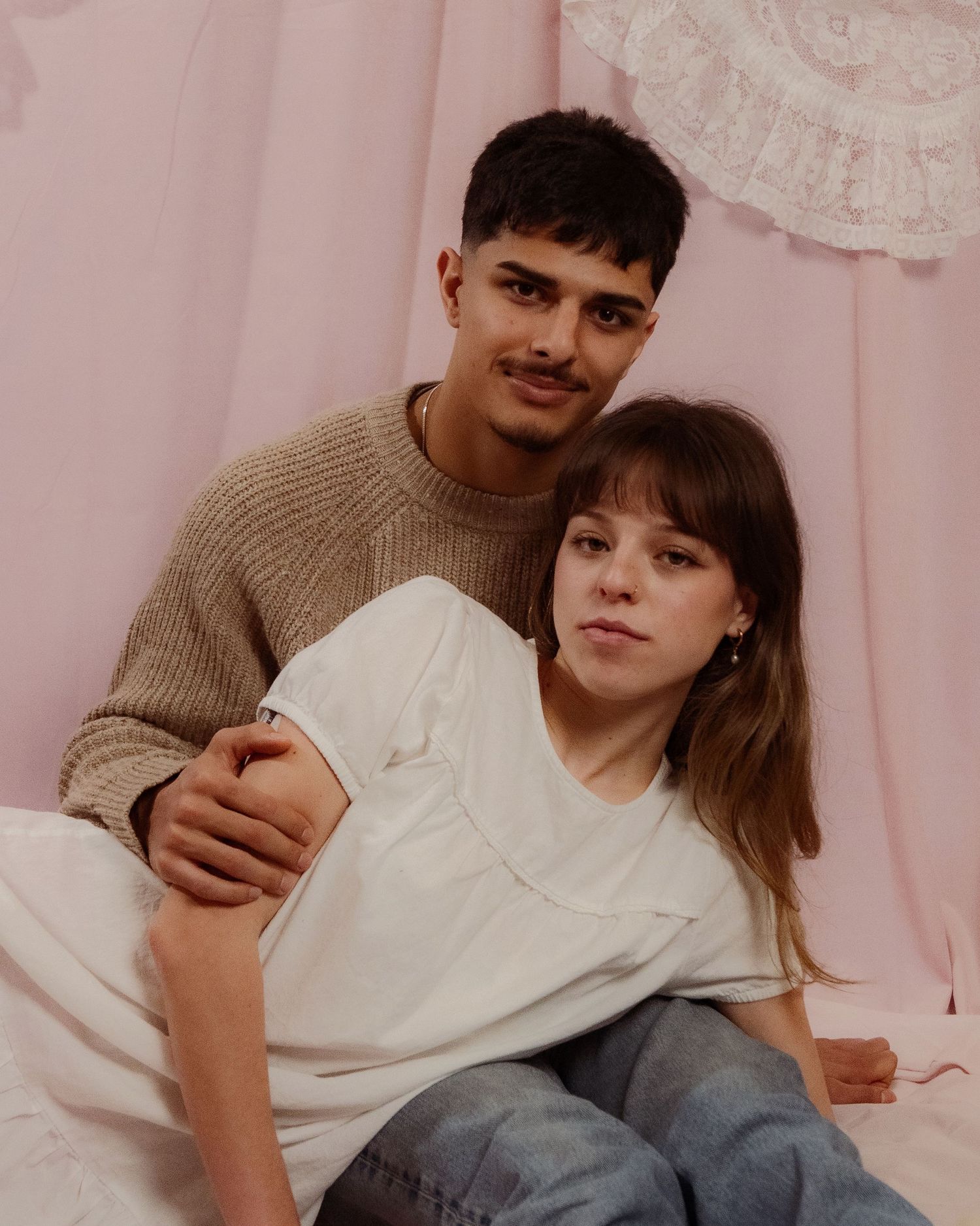 Young couple sitting closely together against a pink backdrop, man in knit sweater holding woman.