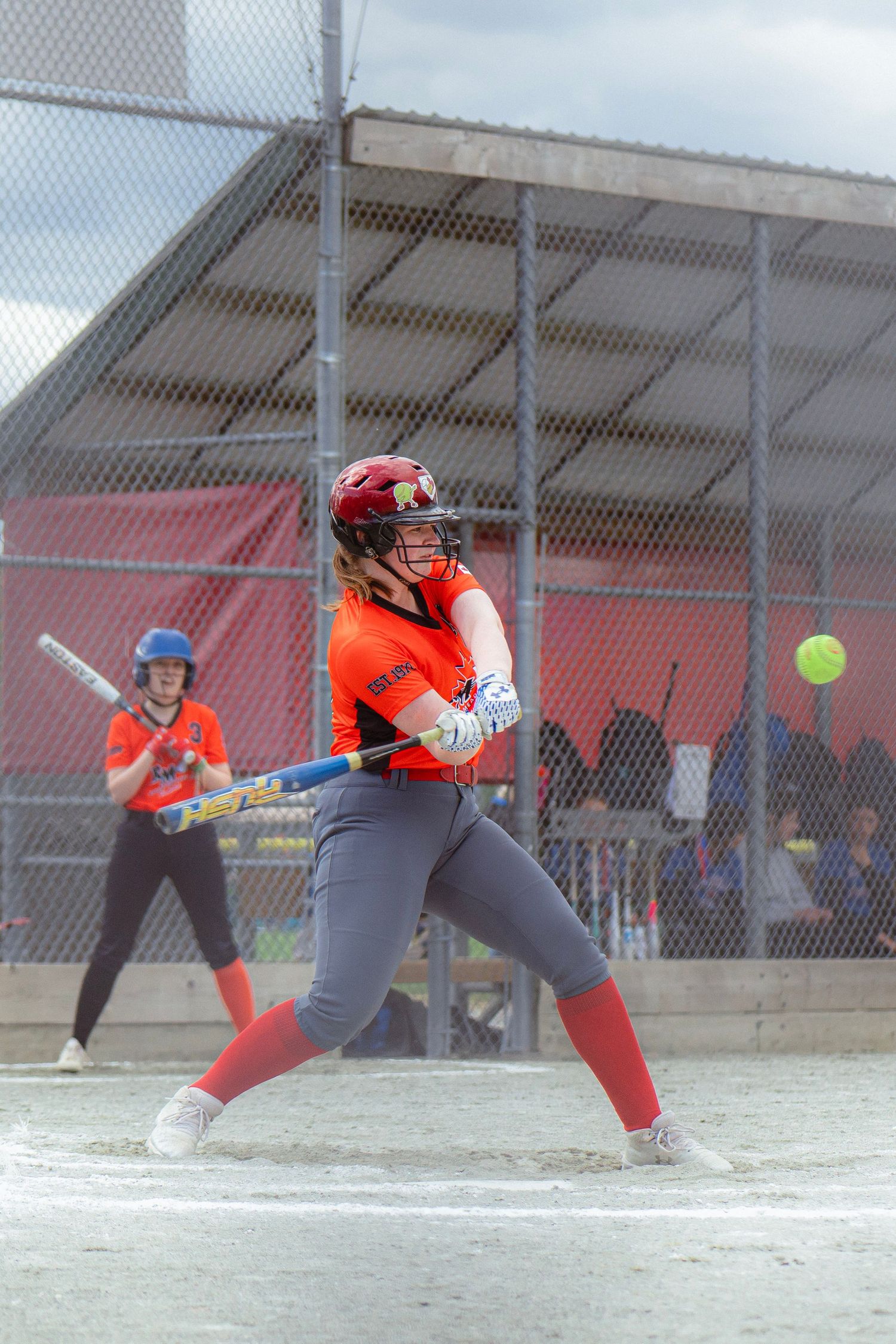 Female softball batter in orange uniform makes contact with incoming yellow ball during game.