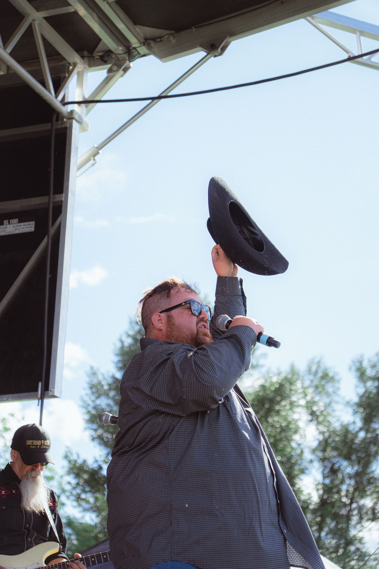 Country singer lifting cowboy hat while performing on outdoor stage at music festival.
