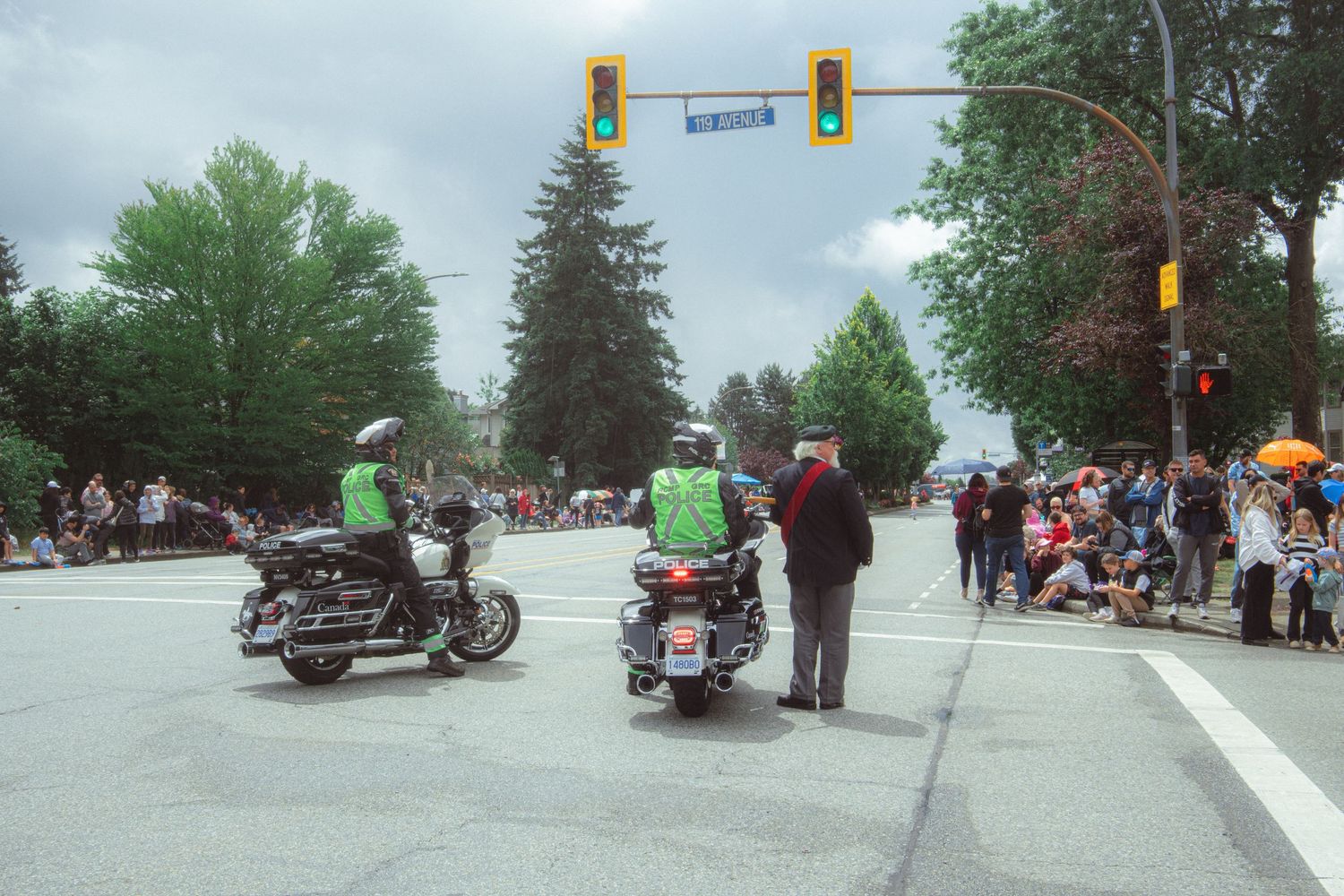 Two green-jacketed motorcycle officers escort a parade past spectators lining a tree-lined suburban street.