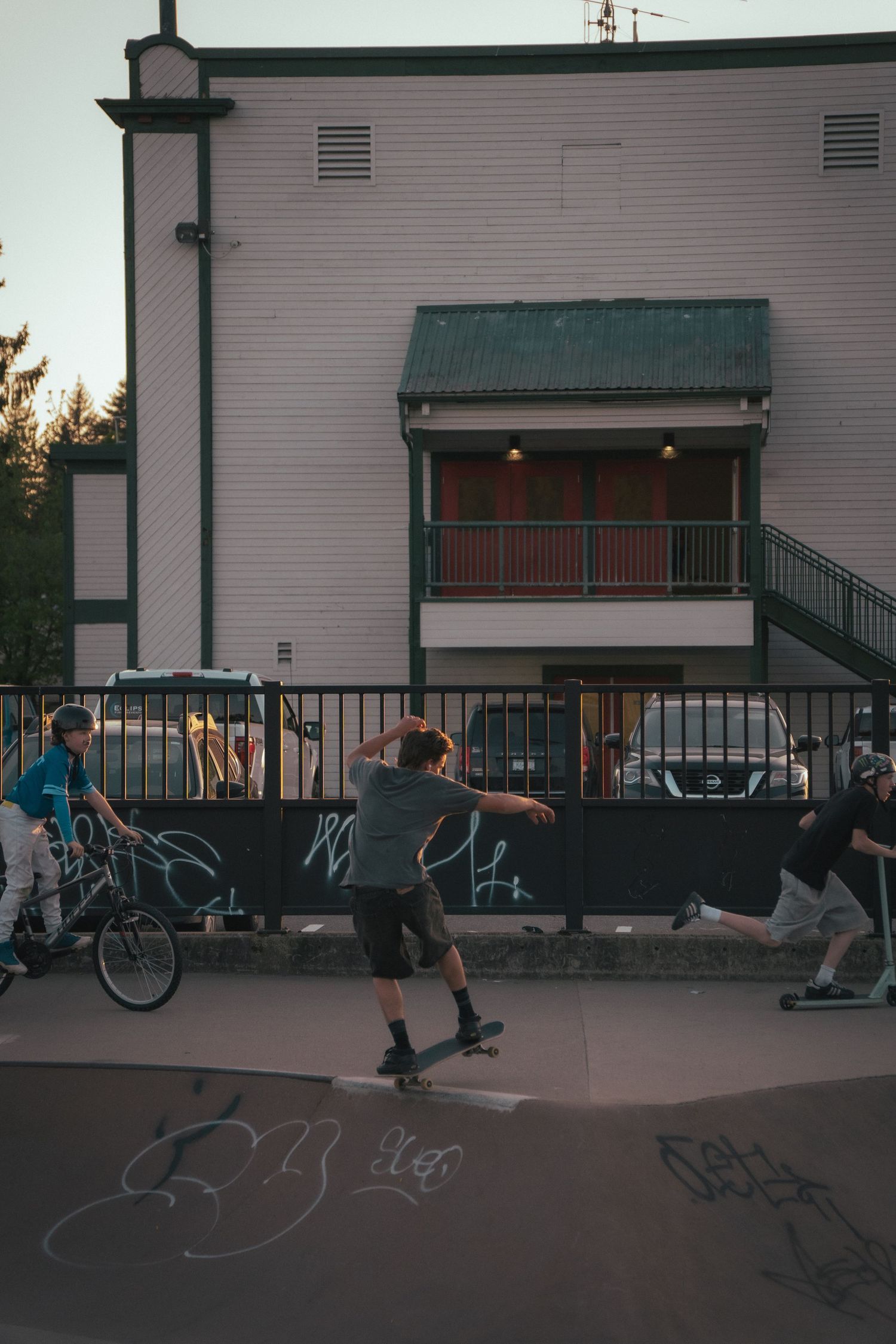 Skateboarders performing tricks at an outdoor skate park near a building with graffiti at dusk.