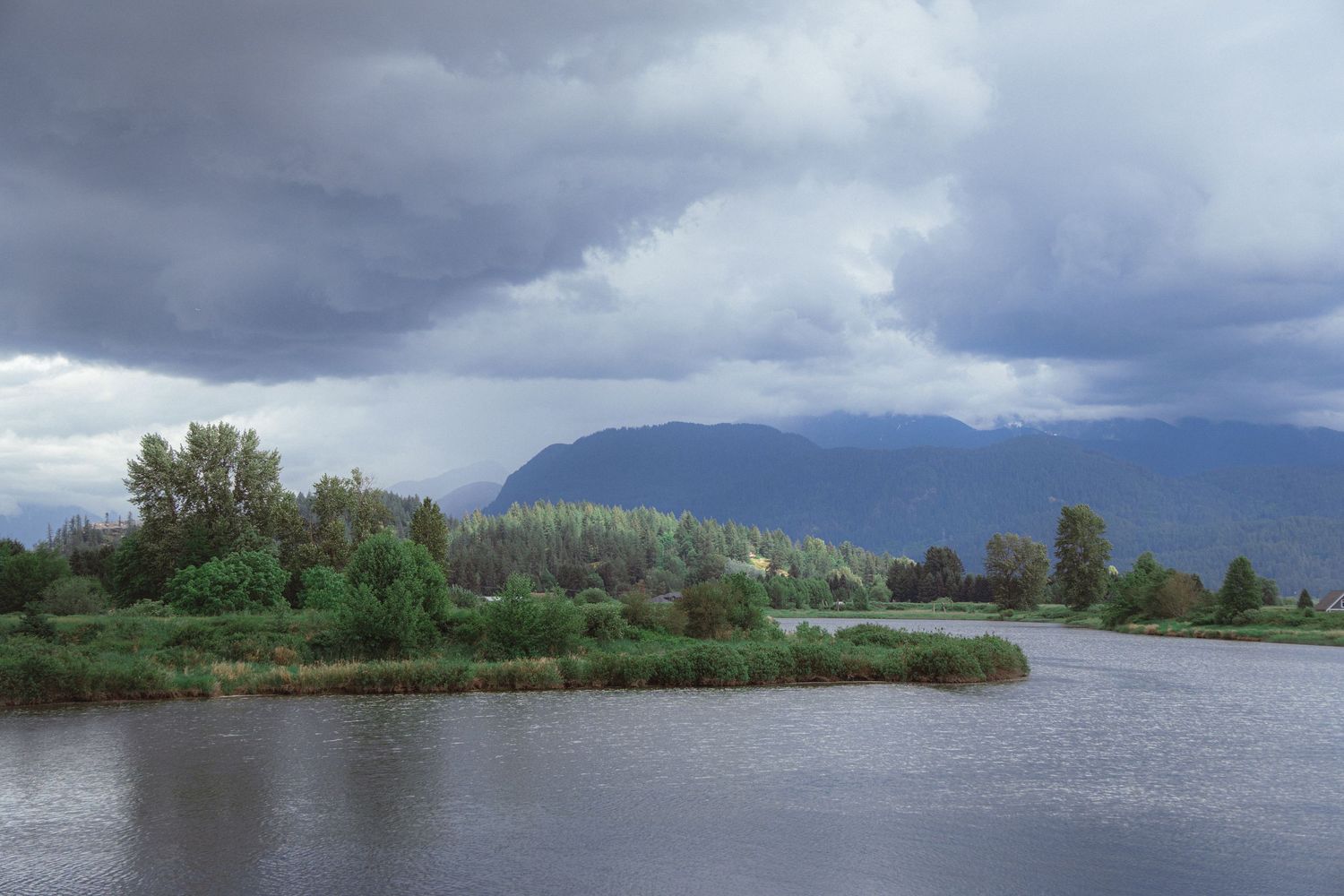 Stormy clouds over a calm lake with lush green trees and mountain backdrop.