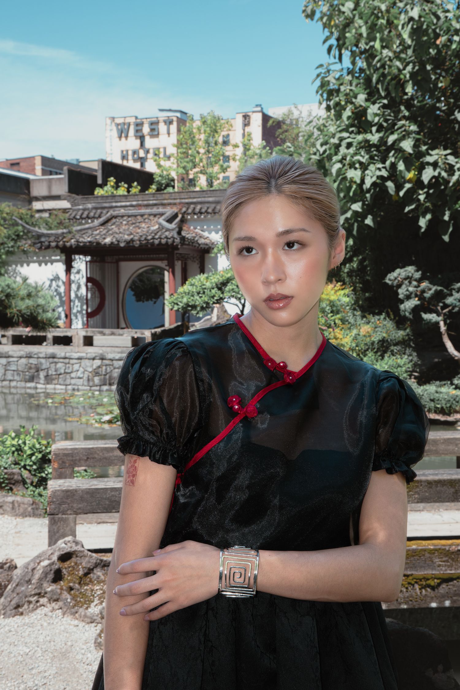 Young woman in black qipao dress with red trim and silver bracelet poses in a Chinese garden.