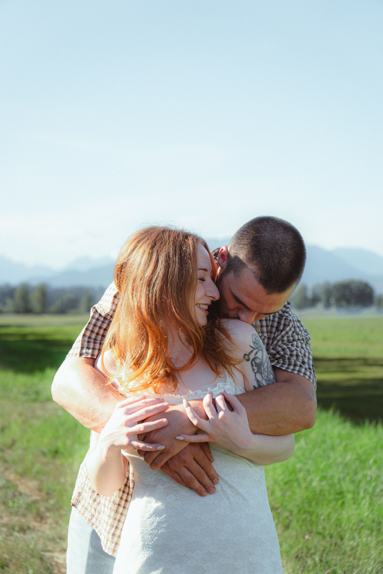 A couple embraces in a green field with mountains, the man hugging the smiling redhead woman from behind.