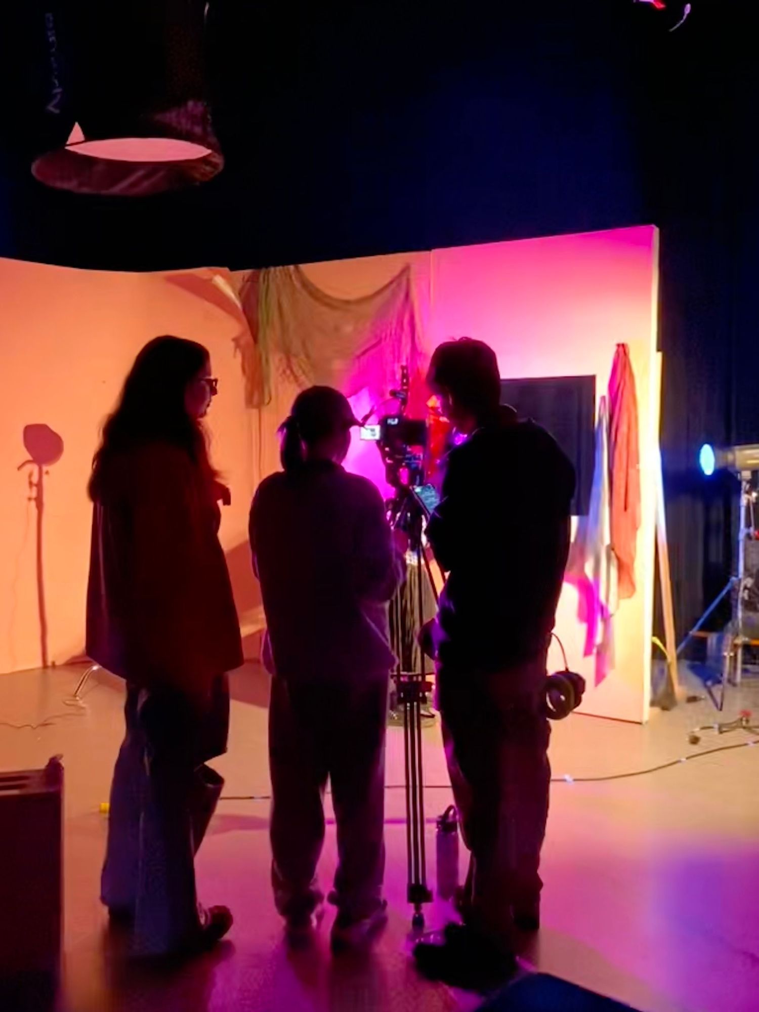 Three filmmakers collaborate around a camera on a tripod in a colorful pink and orange film studio set.