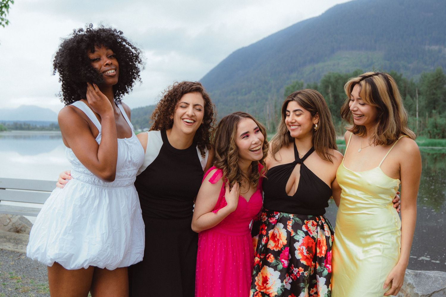 Five diverse women in colorful dresses laughing together with a mountain lake backdrop.
