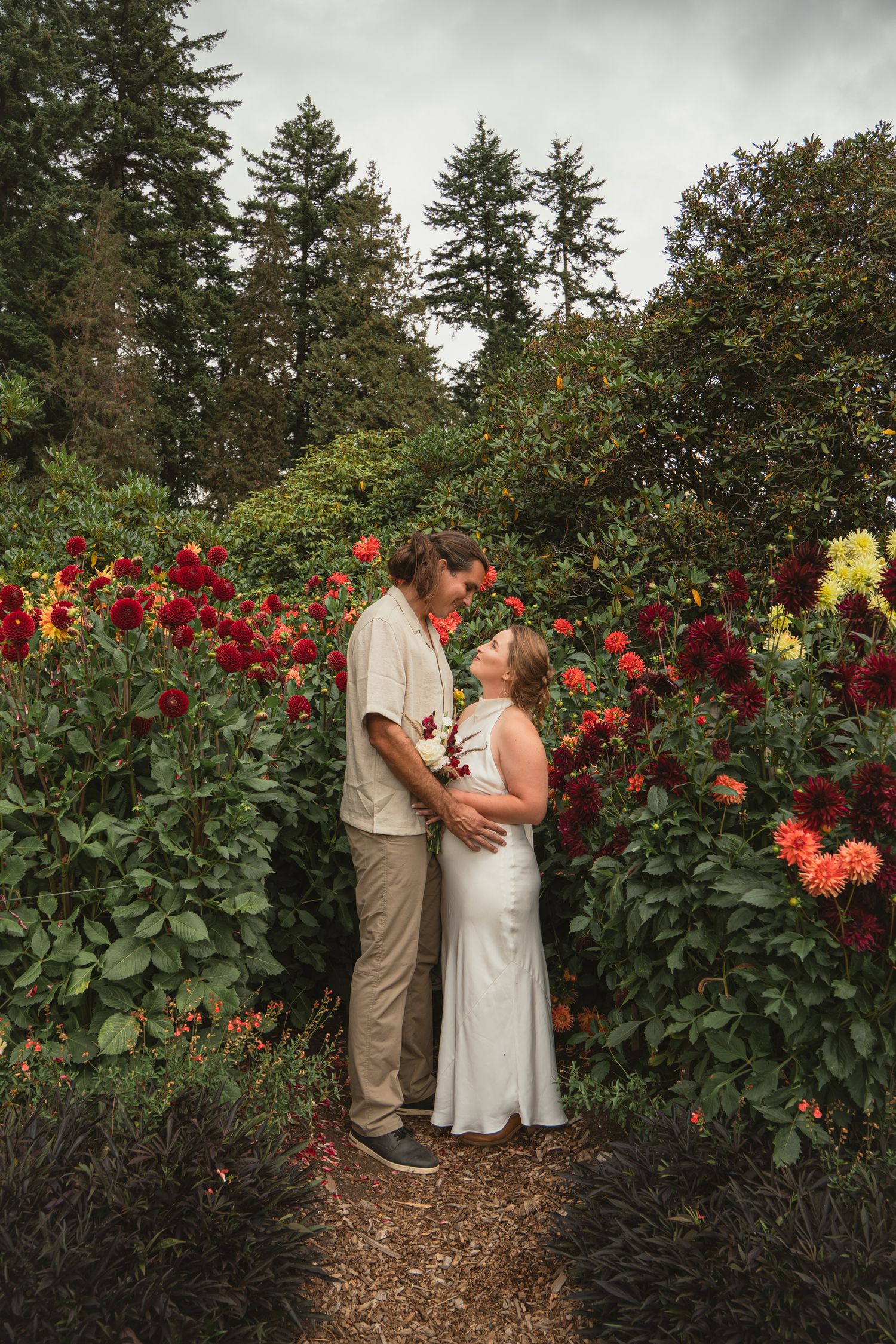 Couple sharing a romantic moment surrounded by vibrant red dahlias in a lush garden setting.