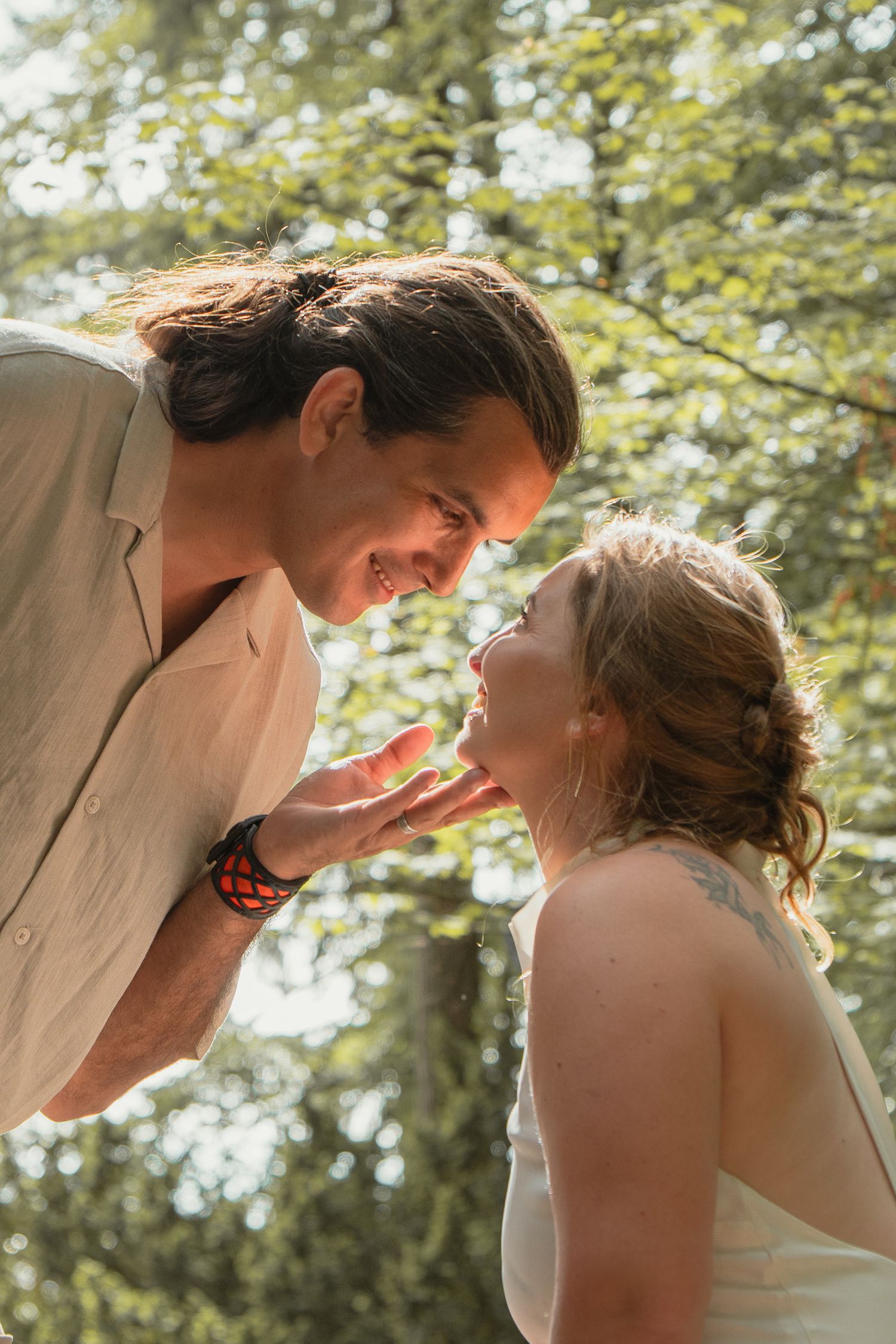Joyful newlyweds sharing an intimate moment, smiling face-to-face in a sun-dappled outdoor setting.