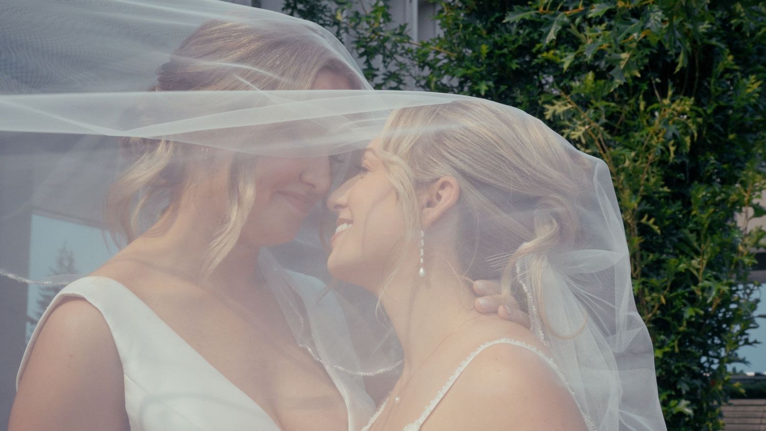 Two brides share an intimate moment under a flowing white veil outdoors on their wedding day.