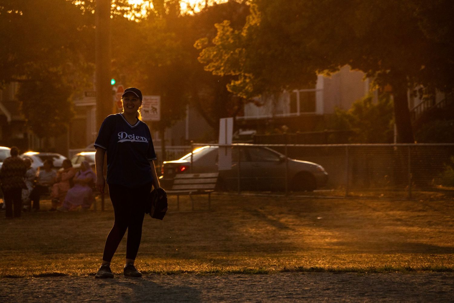 Softball player in Dodgers uniform stands alone on a field bathed in dramatic golden sunset light.