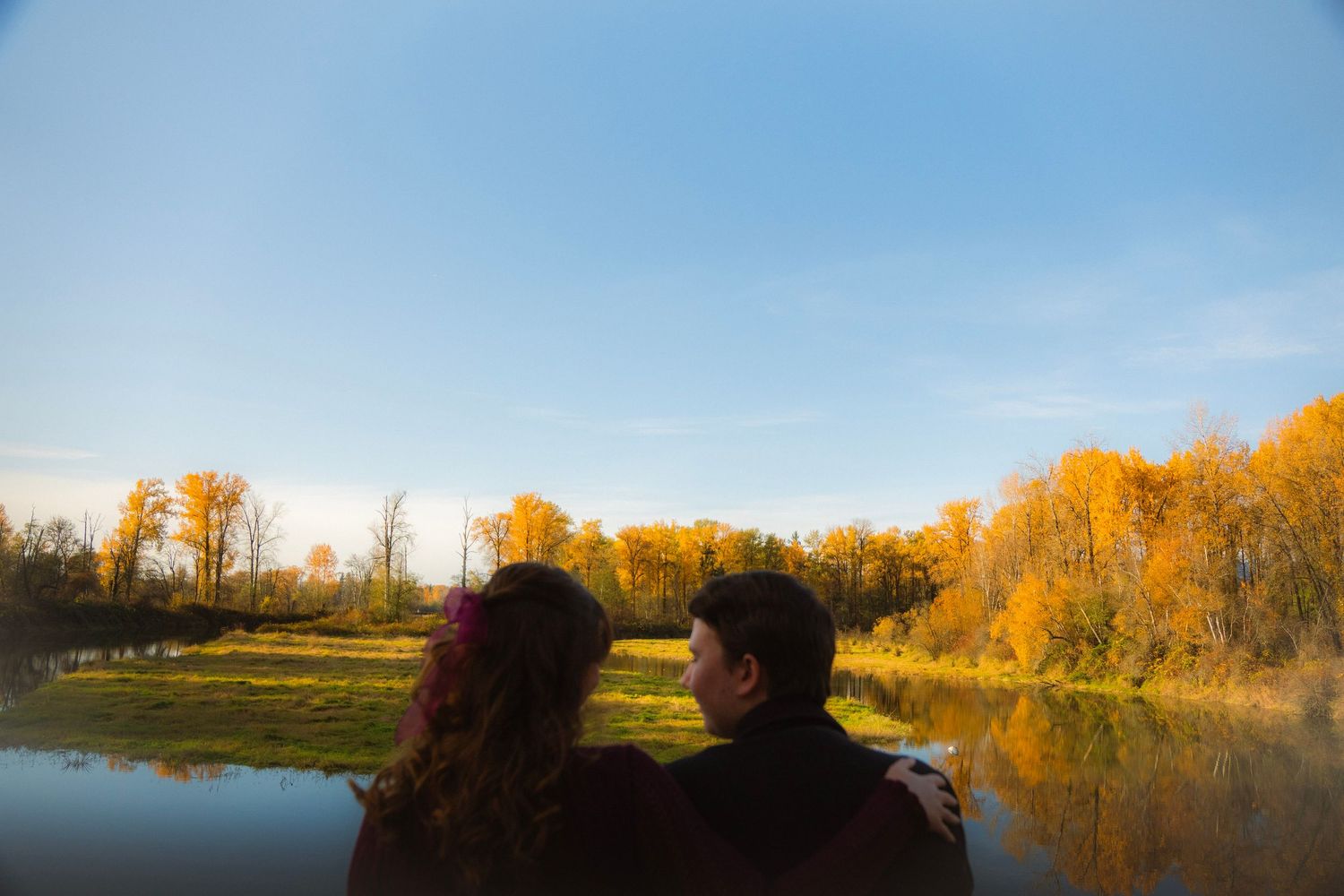 Couple sitting together admiring a golden autumn lake landscape with vibrant yellow trees reflected in calm water.