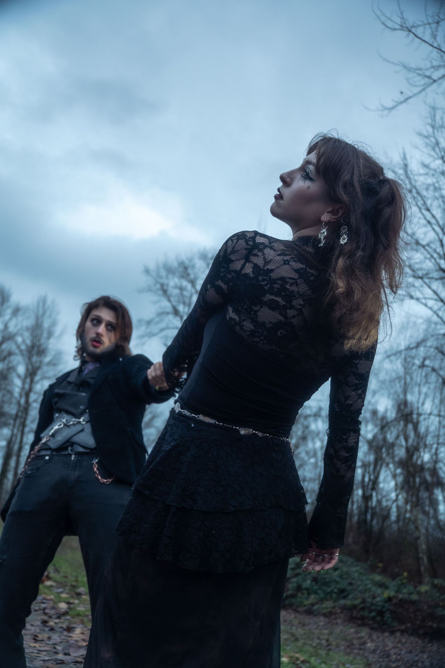 Gothic couple in black lace outfits posing dramatically in a misty forest under an overcast sky.