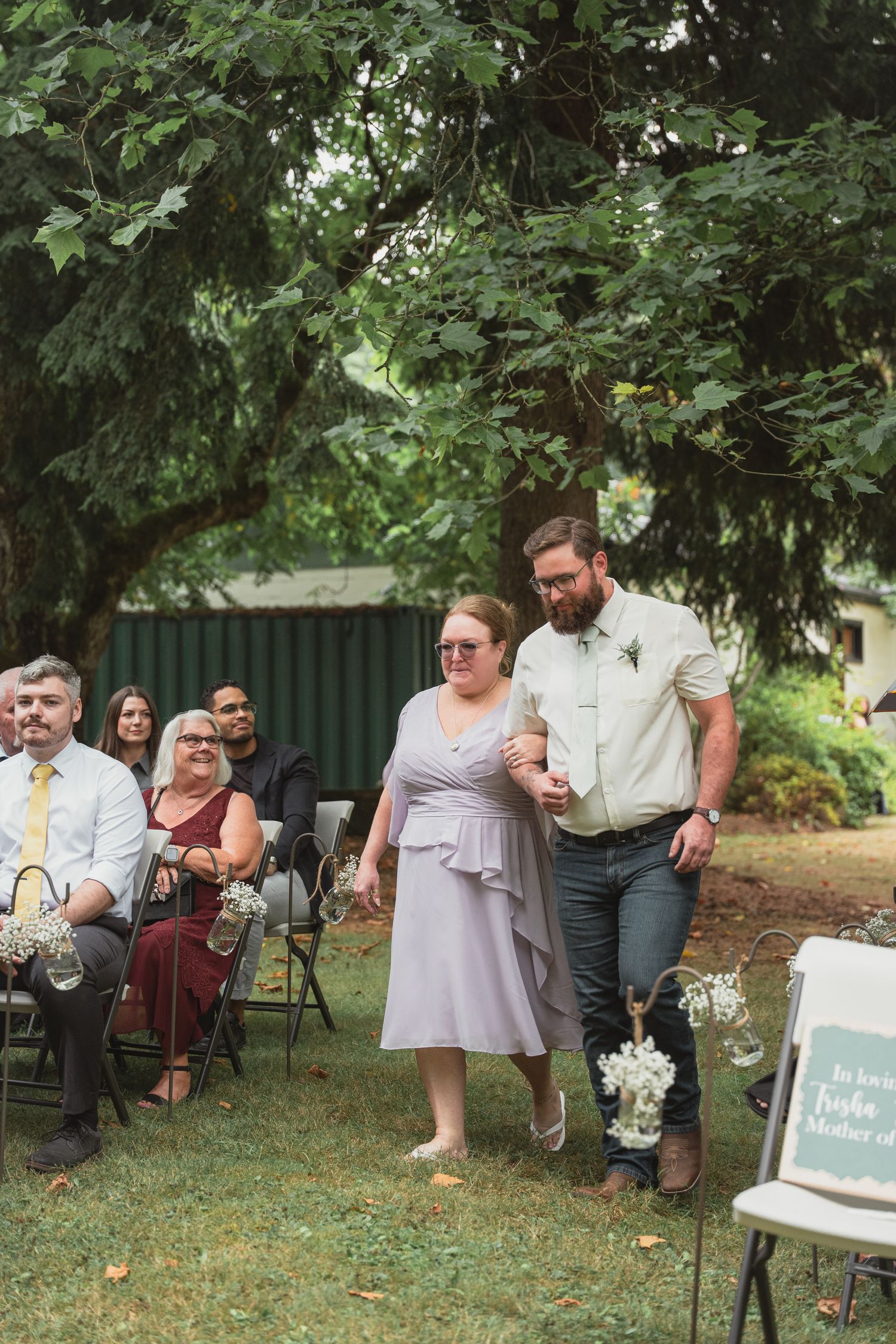 Woman in lavender dress walks down outdoor wedding aisle escorted by bearded man in green shirt.