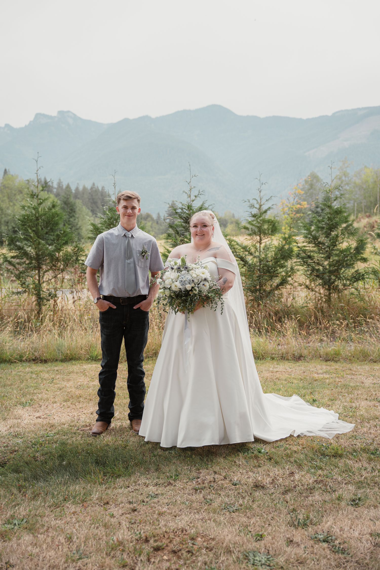 Young man in gray shirt poses beside bride in white gown holding floral bouquet outdoors.