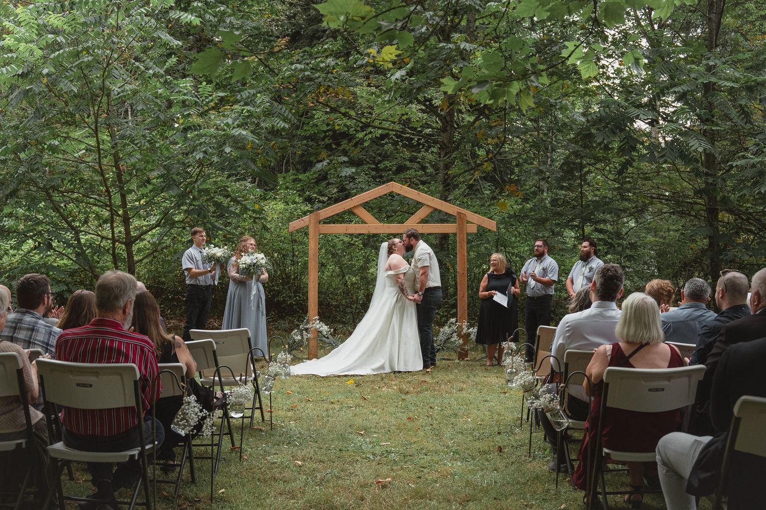 Bride and groom kiss under wooden arch at intimate outdoor woodland wedding ceremony with seated guests.