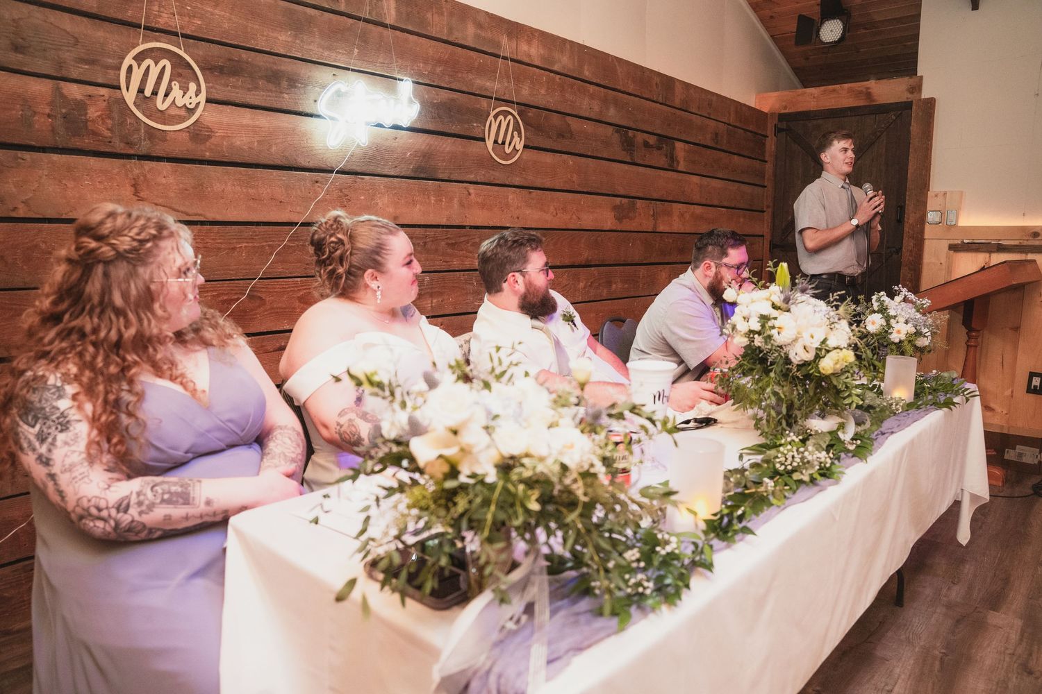 Bridal party listens to groomsman speech at elegantly decorated head table in rustic barn venue.