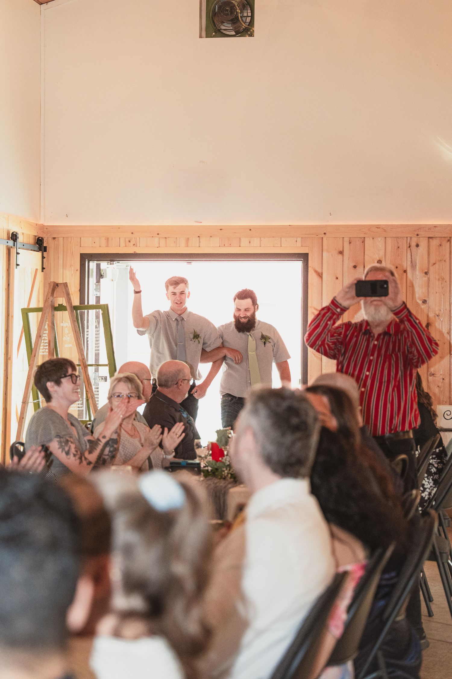 Groomsmen wave to wedding guests seated at tables inside a rustic barn reception venue.