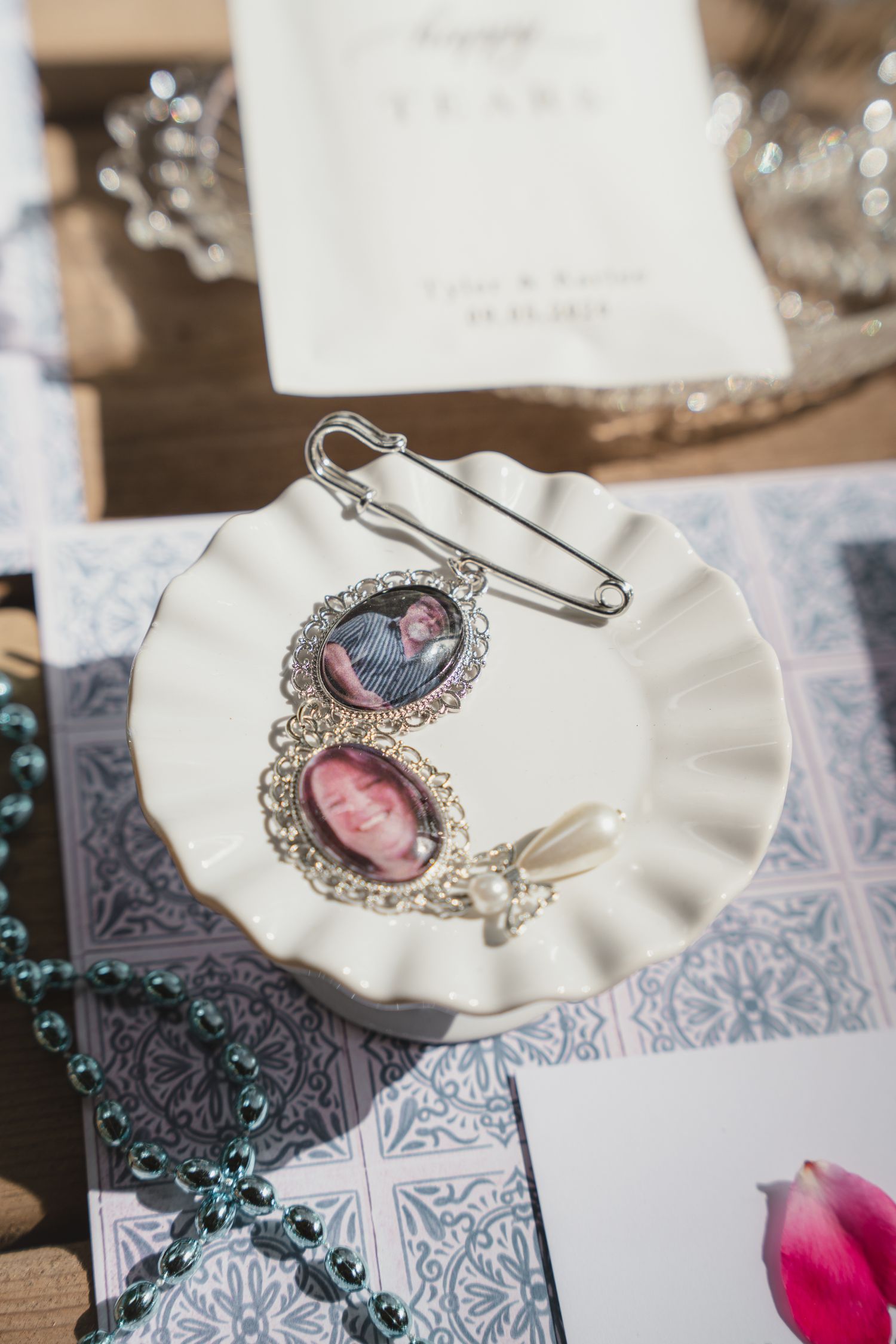 Silver brooch with oval photo lockets displayed on a white ceramic dish alongside pearl beads and wedding stationery.