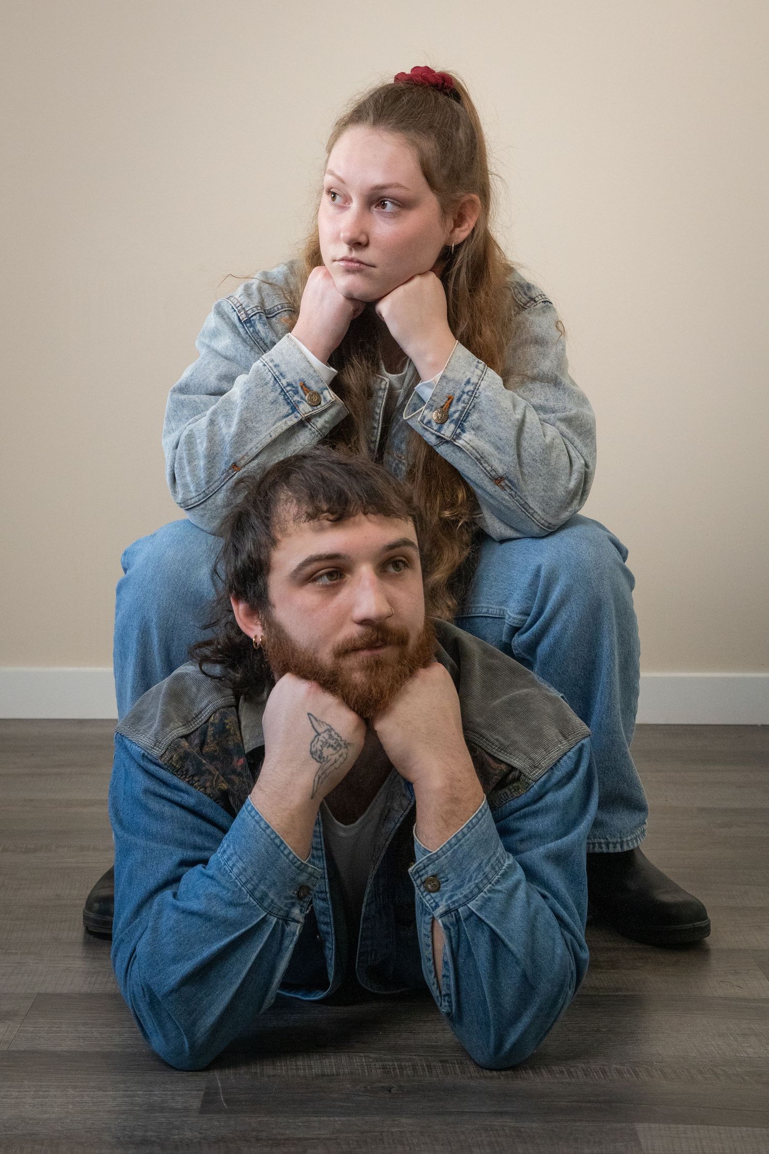 A young couple in matching denim jackets sit on a floor, both resting chins on fists in a bored pose.