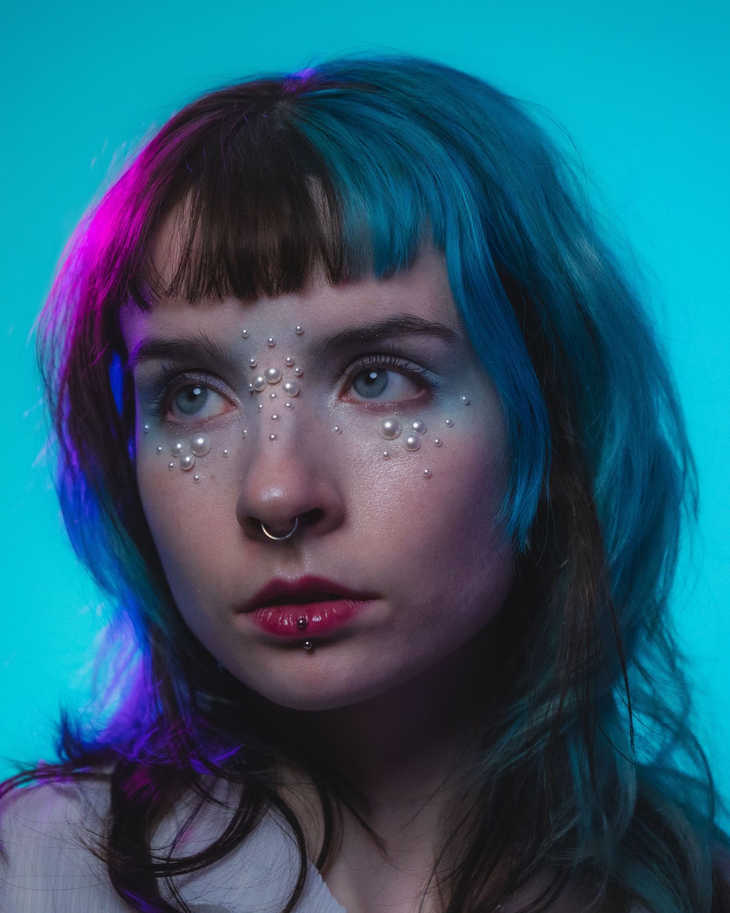 Portrait of woman with pearl face gems, half teal hair, and nose ring under neon blue and pink light.