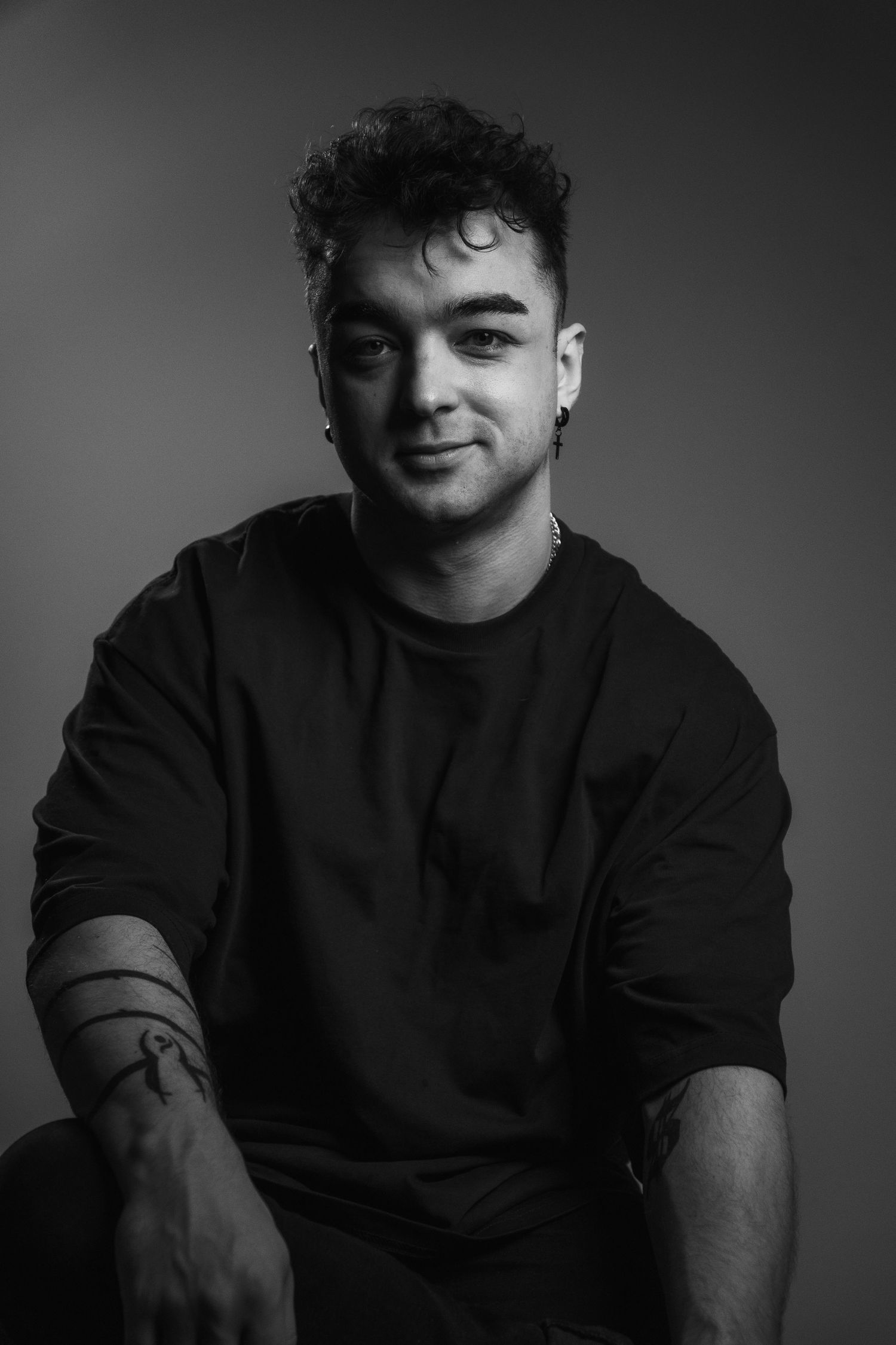 Black and white studio portrait of smiling young man with curly hair, beard, earring, and arm tattoo.
