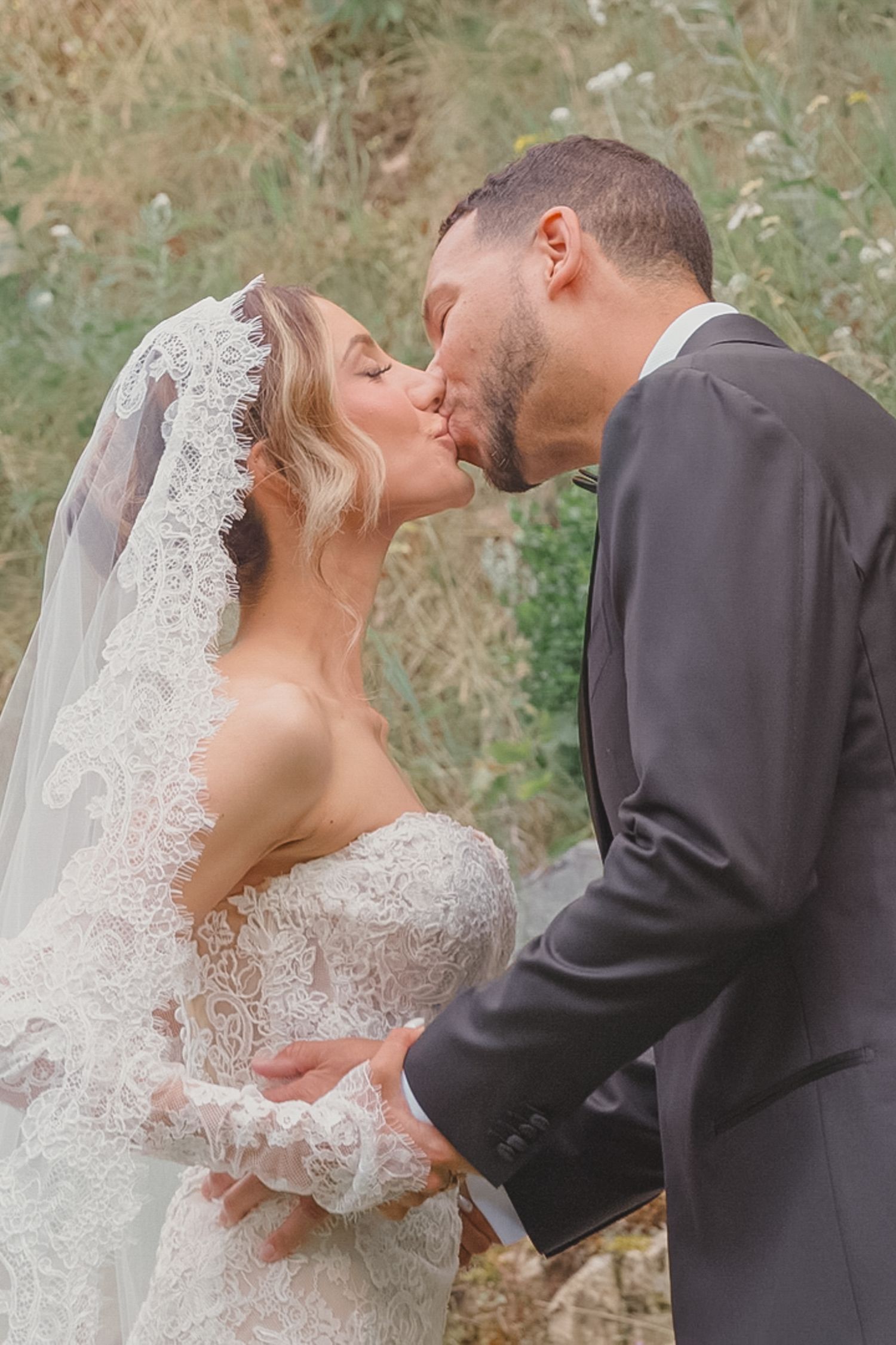 Bride and groom sharing a romantic kiss outdoors, bride wearing lace dress and veil.