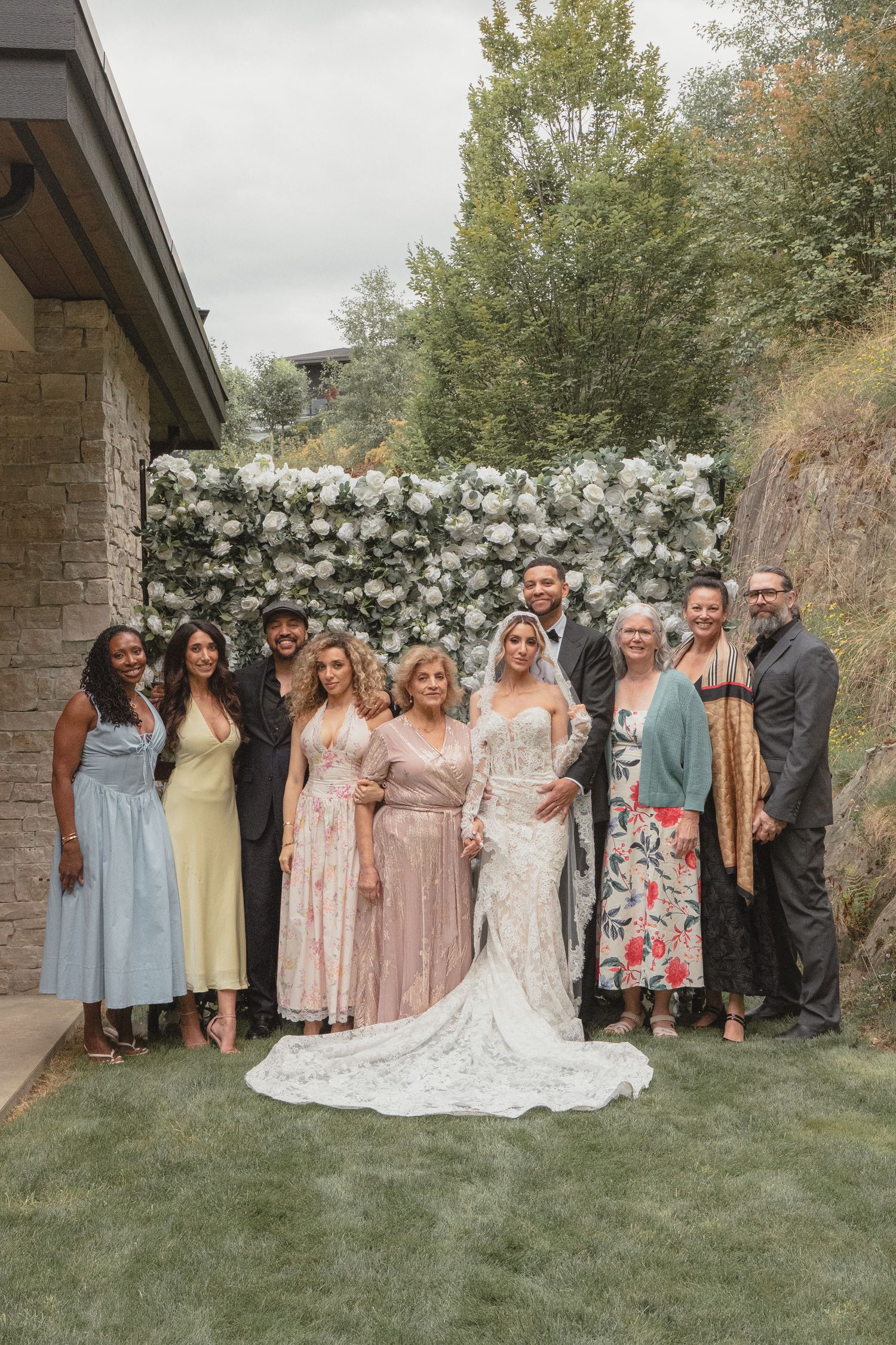Wedding party poses in front of white floral wall backdrop outdoors, bride in lace gown surrounded by family.