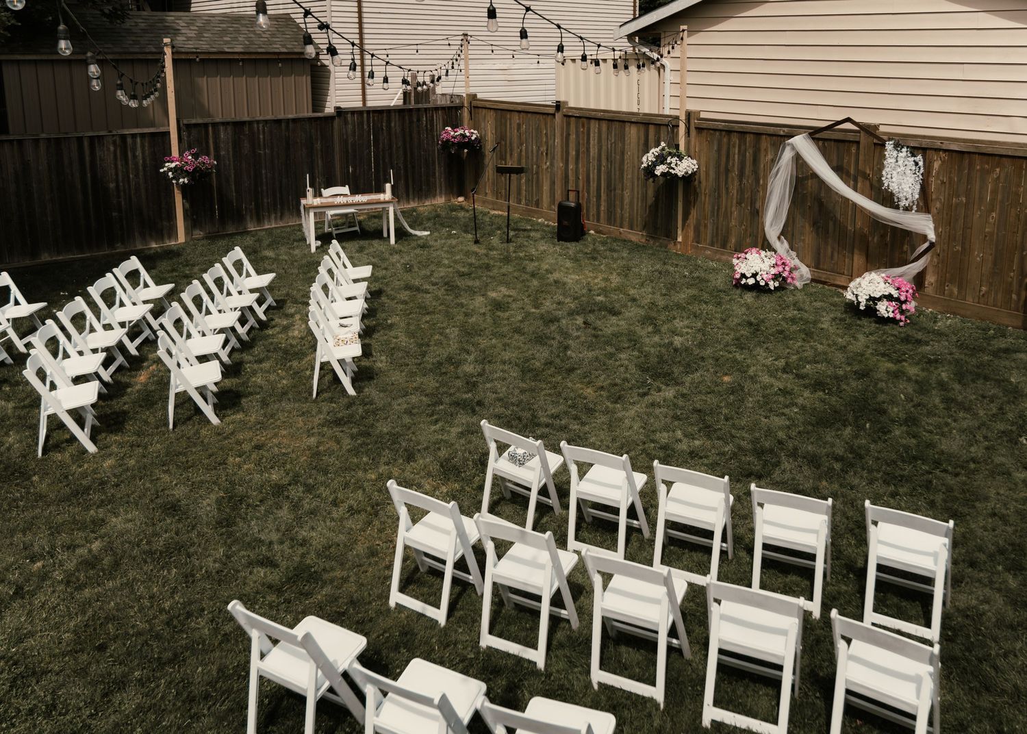Elegant backyard wedding ceremony setup with white folding chairs, floral arch, and string lights on green lawn.