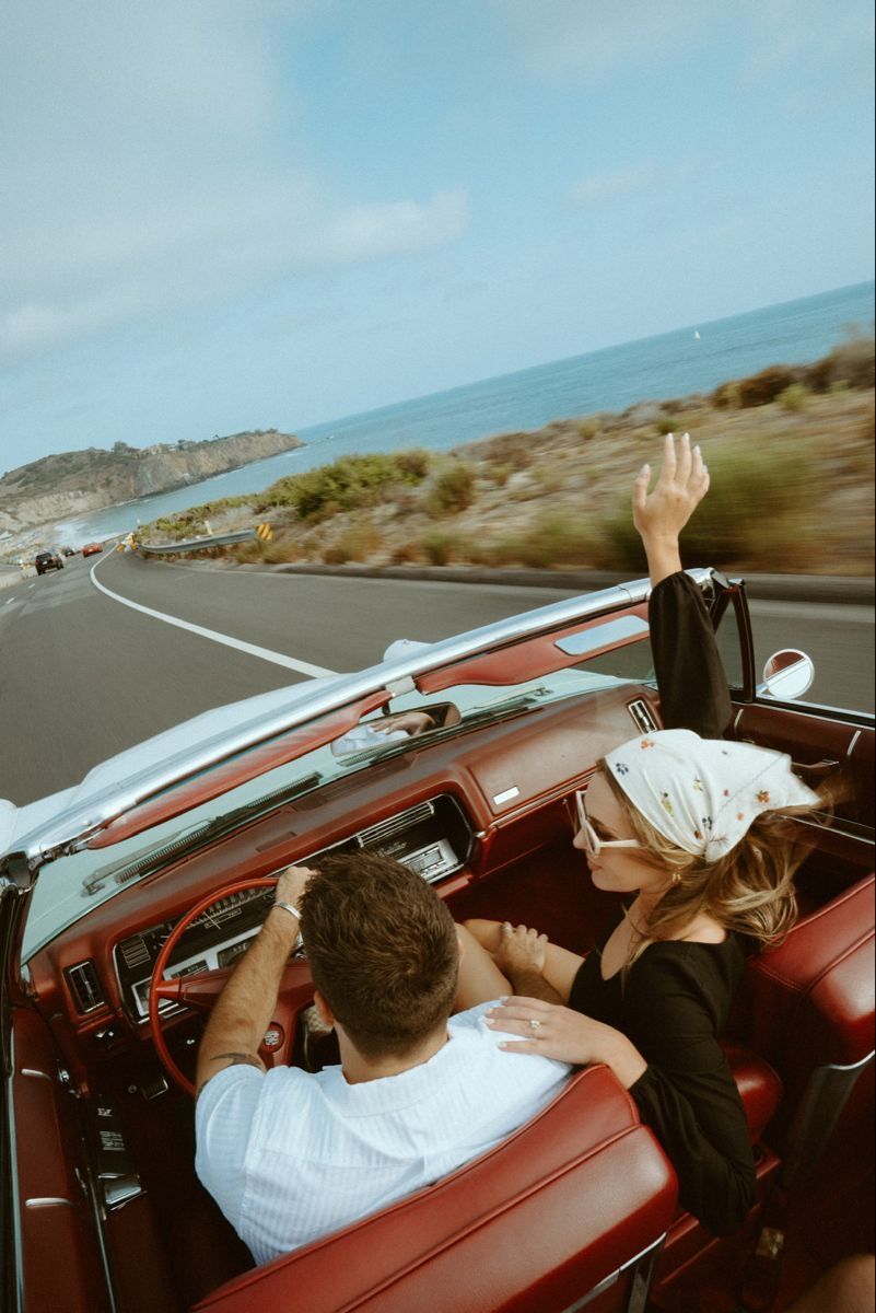Couple driving a classic red convertible along a scenic coastal highway, woman raising hand in the air.