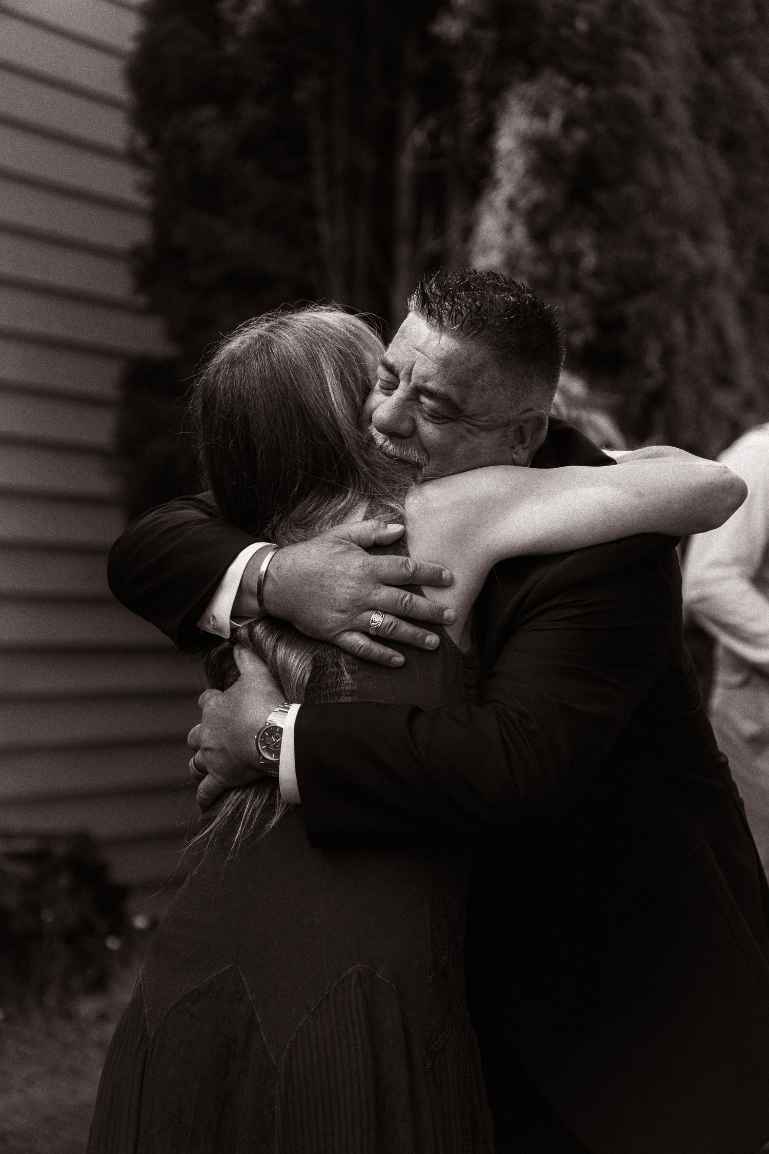 Black and white photo of a man in a tuxedo sharing an emotional hug with a woman at a wedding celebration.