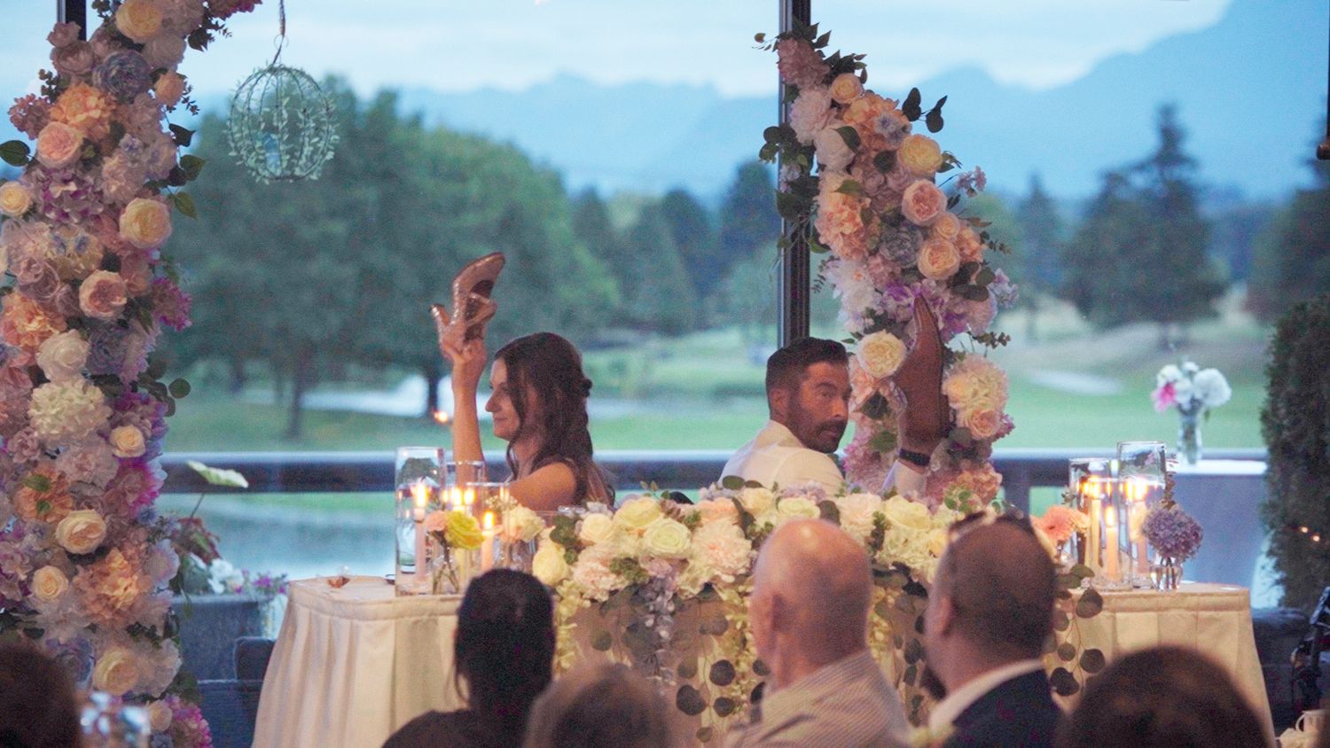Bride and groom seated at floral-adorned sweetheart table during elegant outdoor wedding reception with guests.