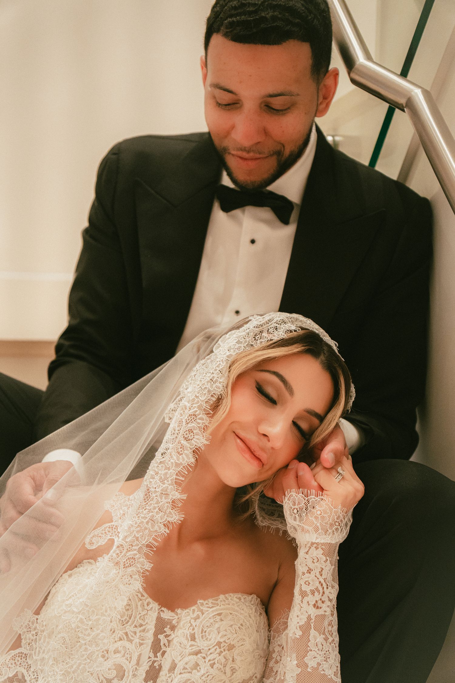 Bride in lace wedding gown and veil rests on groom in black tuxedo on stairs during romantic wedding portrait.