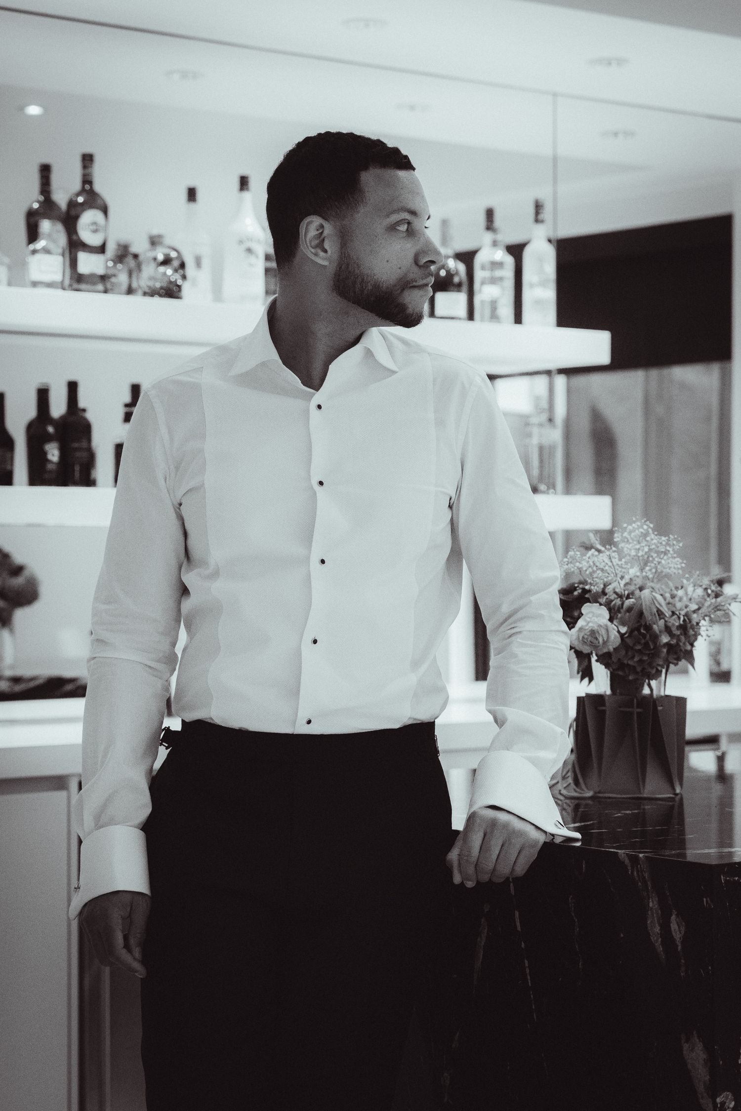 Black and white photo of a well-dressed man in a white dress shirt standing by a bar with bottles on shelves.