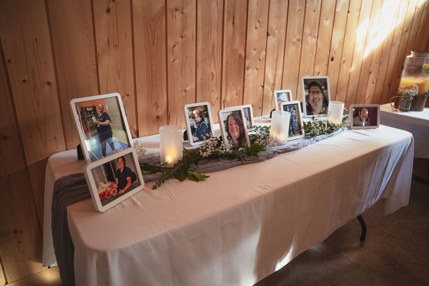 Memorial table with framed photos, greenery, and candles on a white tablecloth in a rustic wood-paneled room.