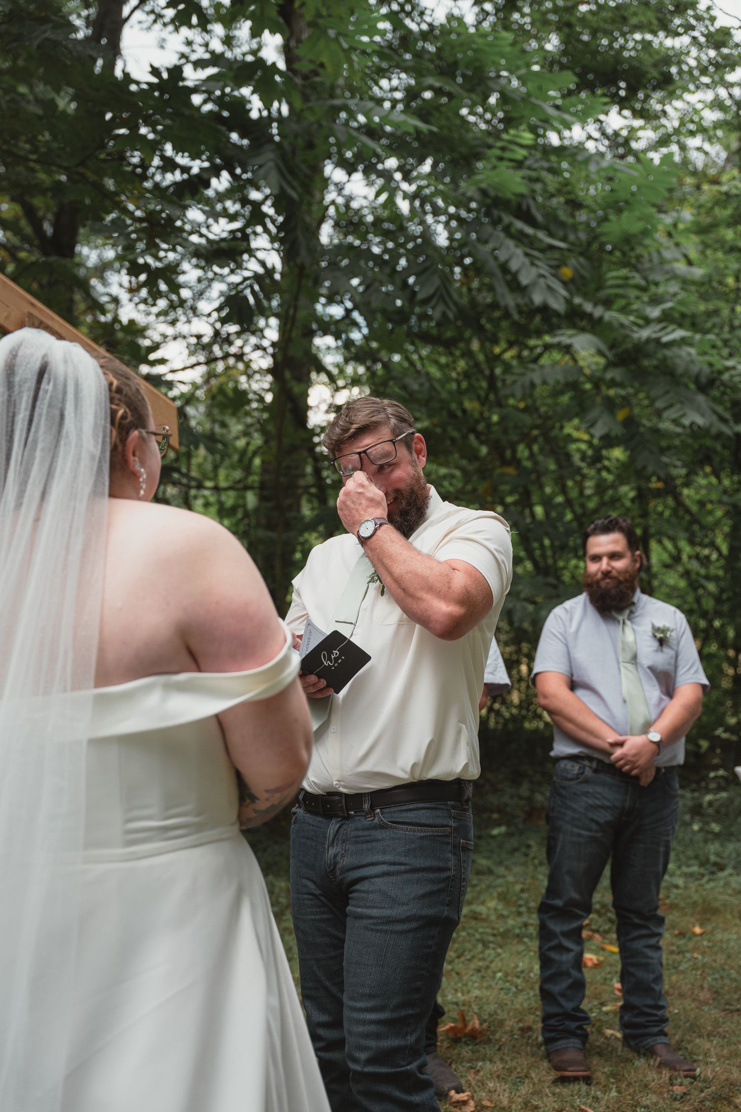 Emotional groom wiping tears during outdoor wedding vows while officiant reads from notes beside bearded groomsman.