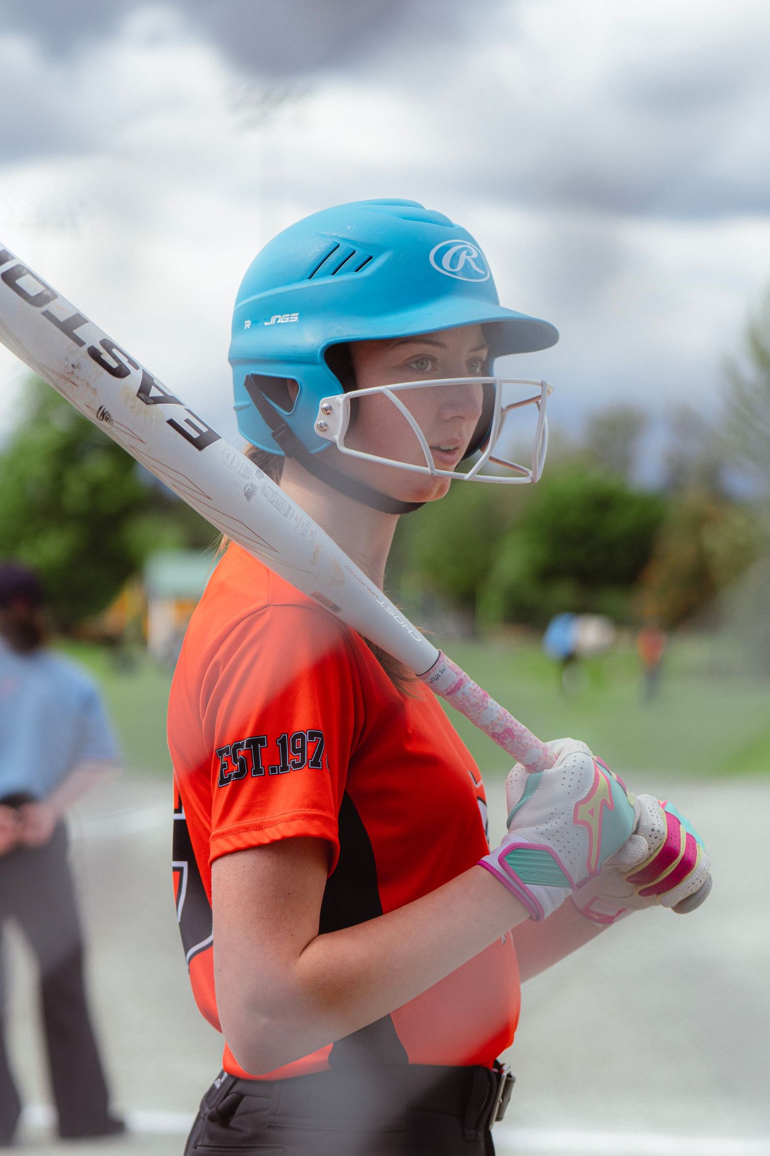 Young female softball batter in blue Rawlings helmet and orange jersey holds Easton bat at plate.