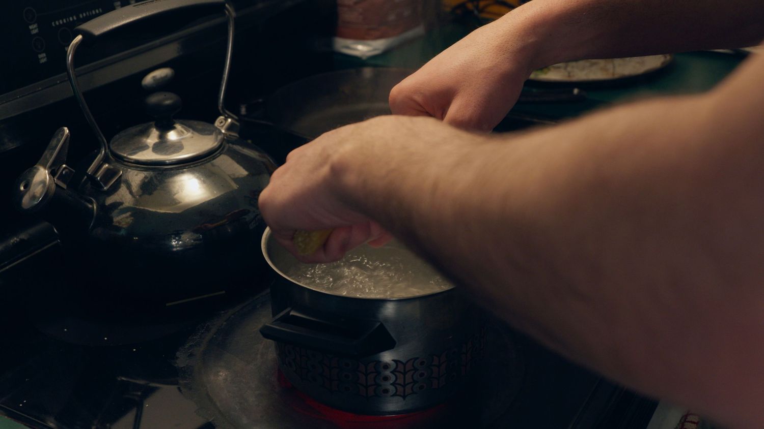 Close-up of hands stirring food in a pot on a dark stovetop beside a vintage black kettle.