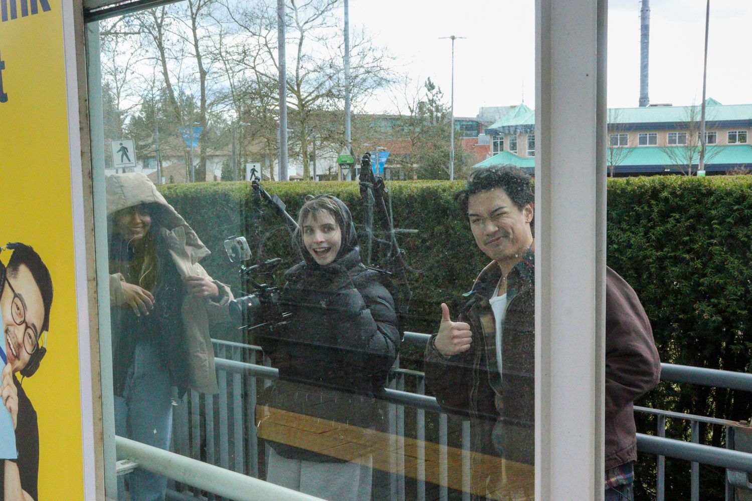 Three film crew members smile through glass at a transit station filming location.