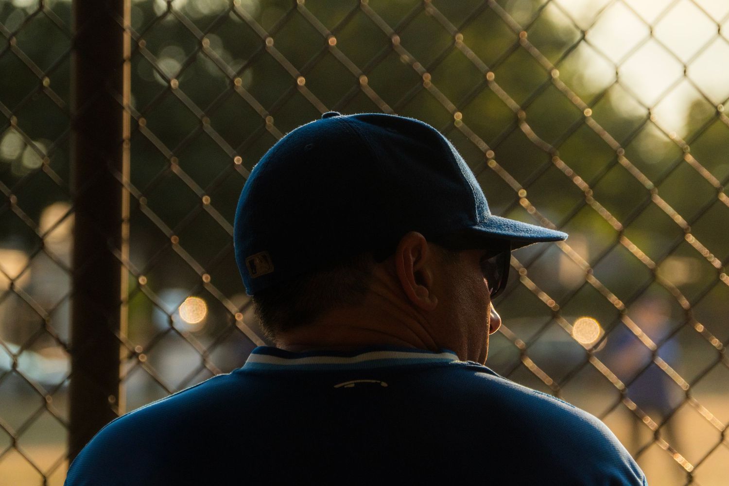 Back view of a man in a blue baseball cap watching a game through a chain-link fence at sunset.