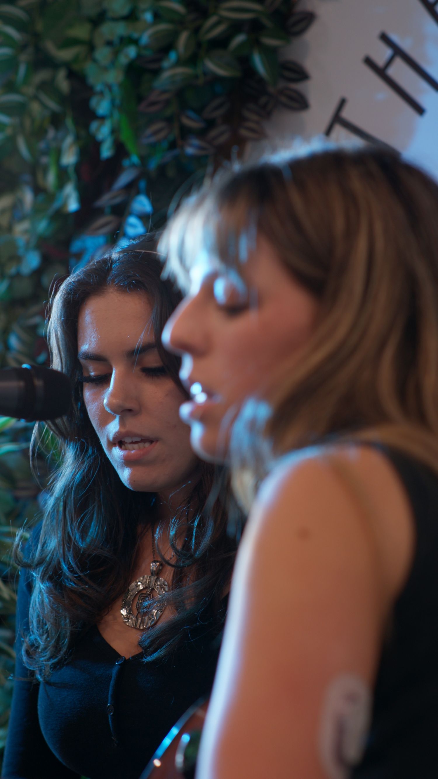 Two women performing live music near microphone surrounded by lush green plants in dimly lit venue.