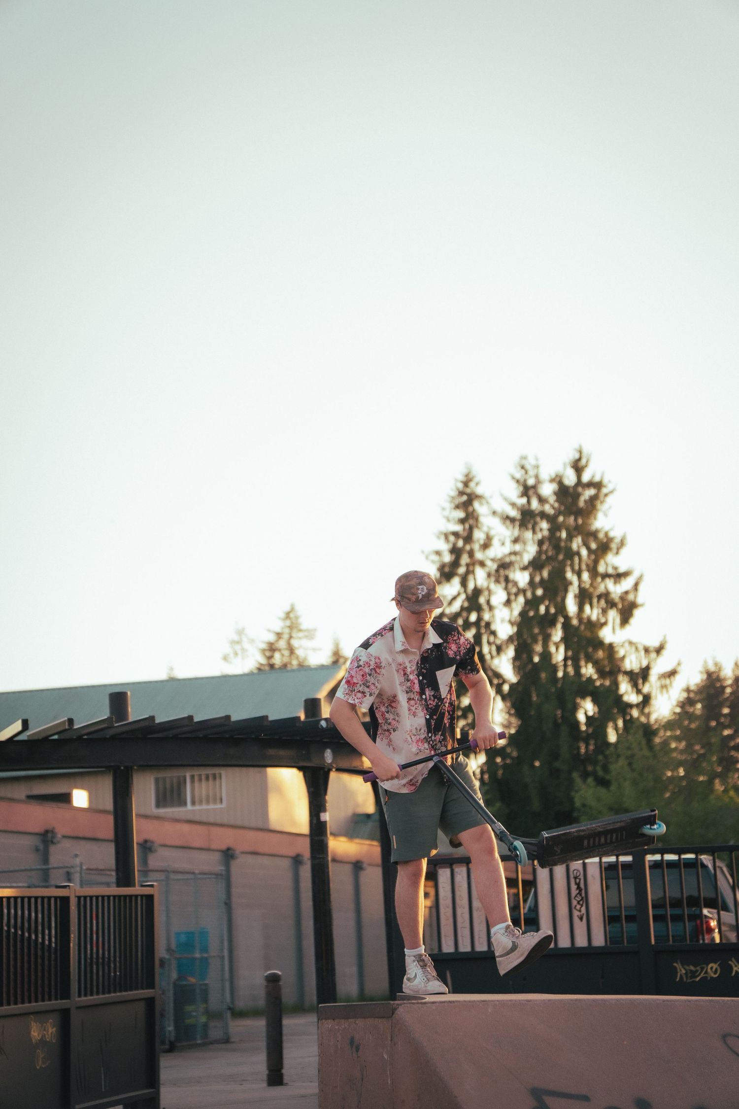 Man in floral shirt holding a scooter while standing on a skate park ramp at dusk.