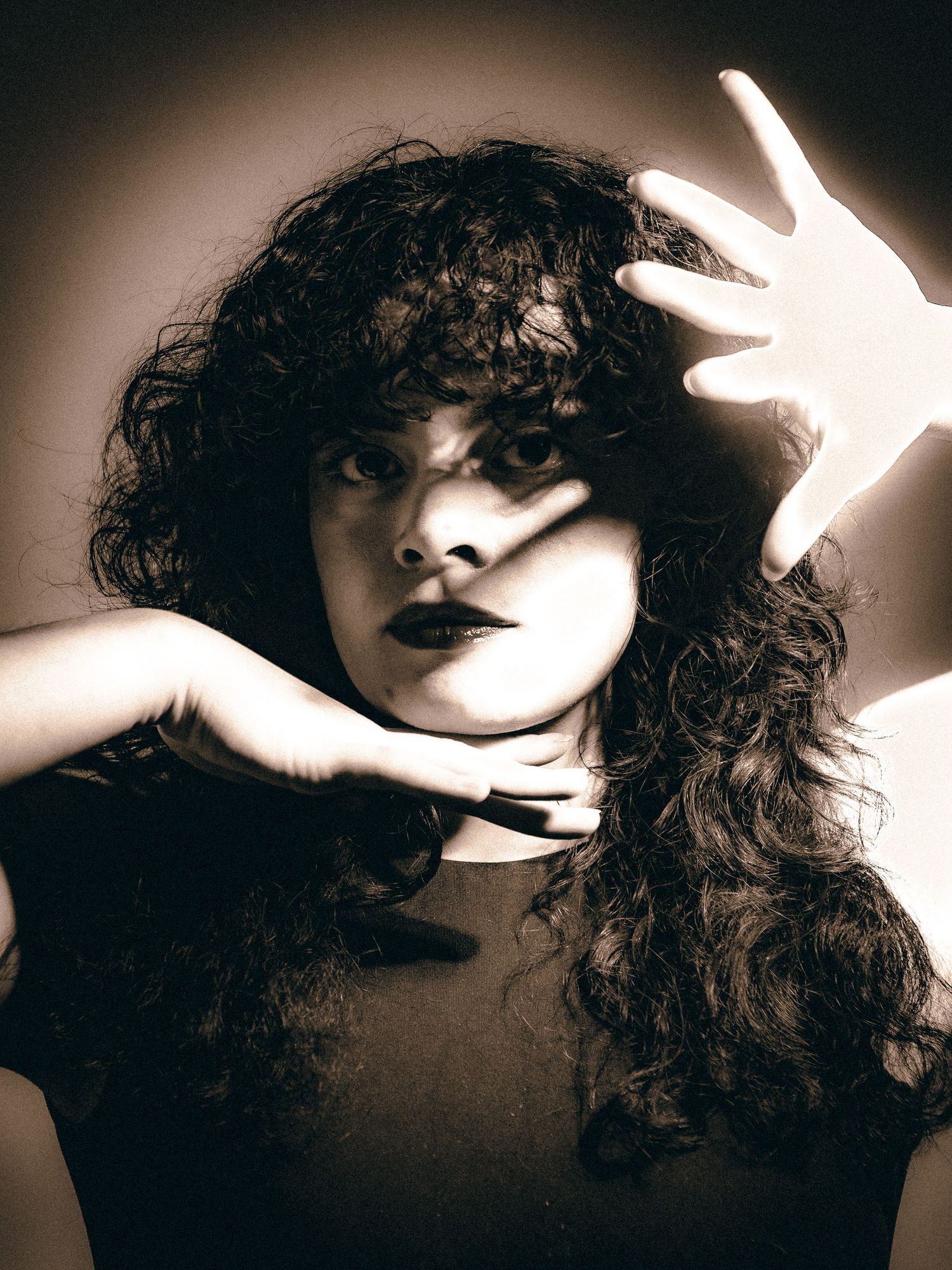 Dramatic sepia portrait of curly-haired woman with hands raised in a defensive or expressive pose.