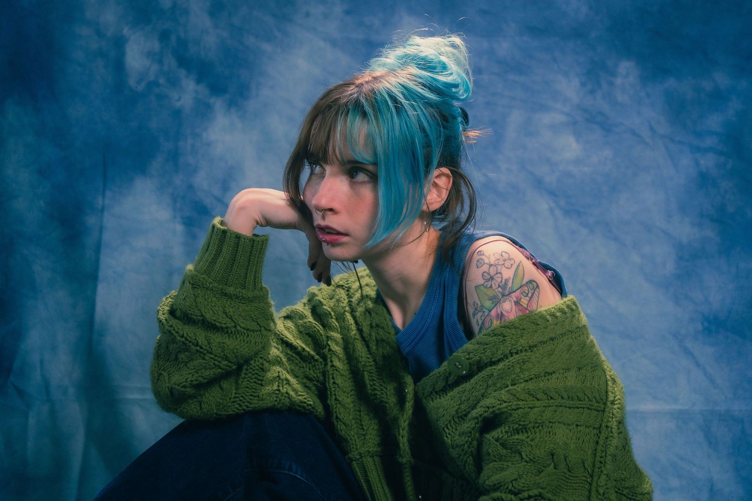 Close-up of woman with teal hair and arm tattoo wearing green sweater posing in blue-toned studio.