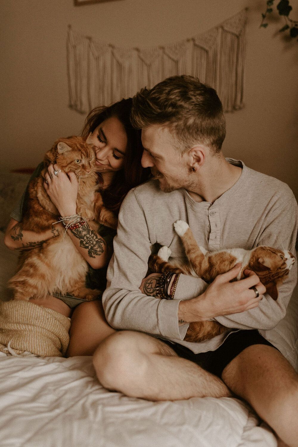 Couple sitting on bed holding cats, woman with tattoos nuzzling orange tabby, man kissing small kitten in cozy boho bedroom.
