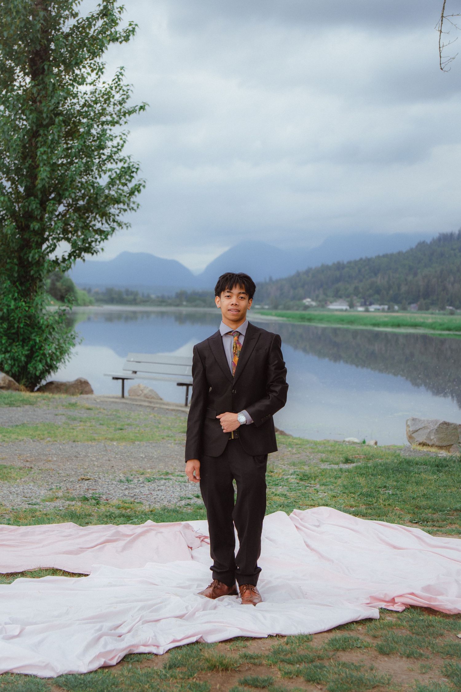Young man in dark suit stands on pink fabric by a misty mountain lake.