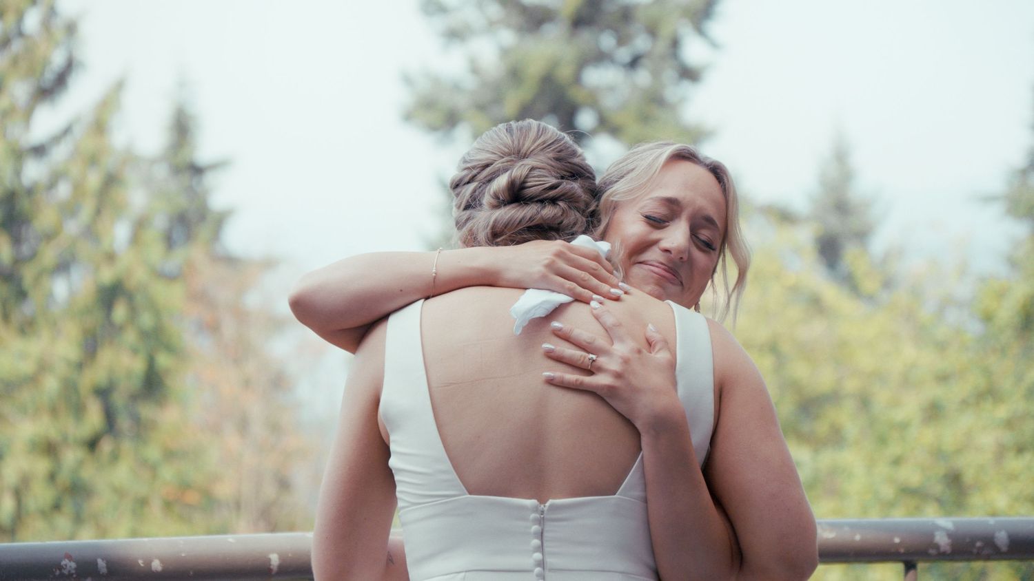 Two brides sharing a heartfelt hug outdoors on their wedding day, surrounded by lush green trees.